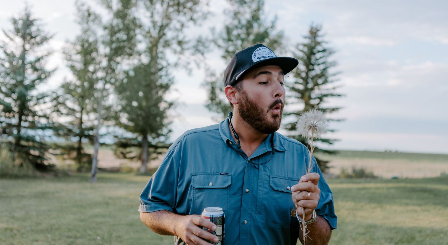 Man in blue shirt and trucker hat holding a drink outdoors in a scenic rural field with trees.