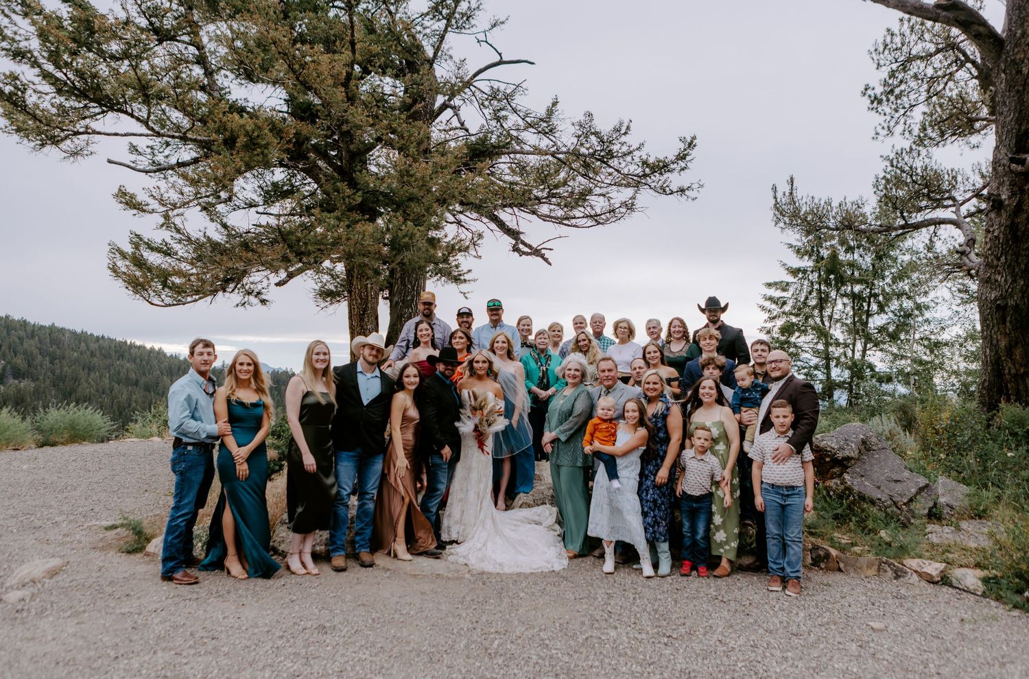 Large wedding party group photo outdoors near pine trees on a gravel overlook with mountain views.