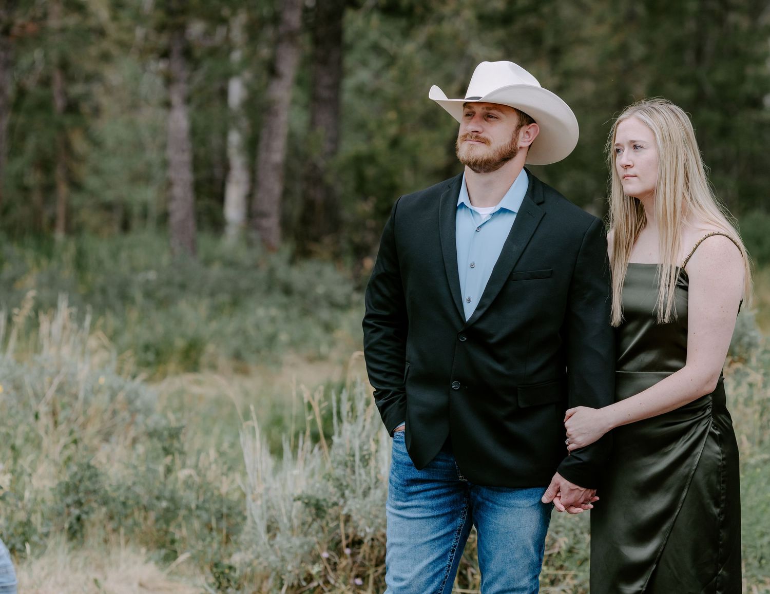 Couple holding hands in a field, man wearing cowboy hat and blazer, woman in green dress, forest background.