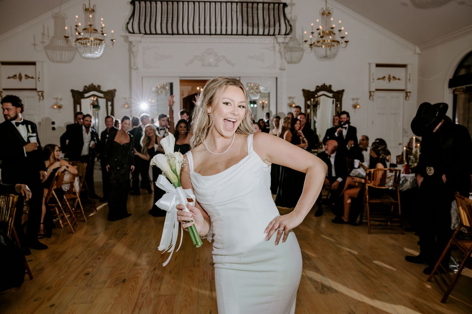 Smiling bride in a fitted white gown holds a calla lily bouquet as wedding guests look on in an elegant ballroom with chandeliers.