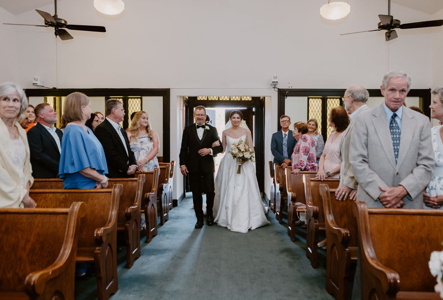 Bride walks down church aisle with escort during wedding ceremony, guests seated in wooden pews on both sides.