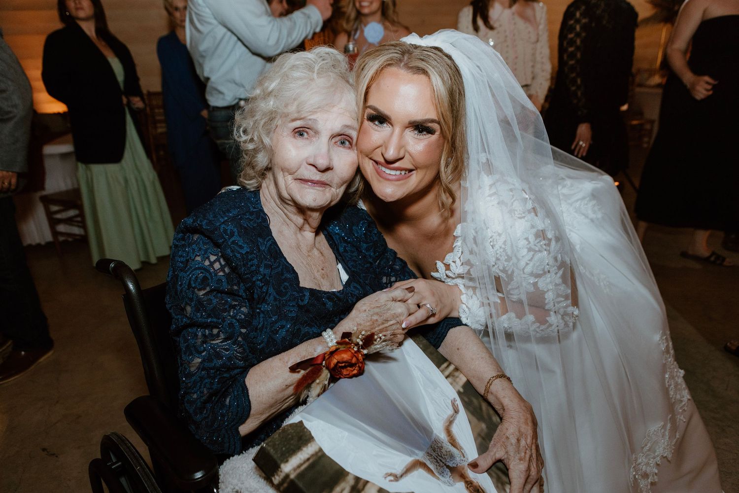 Bride in lace wedding gown poses with elderly grandmother in wheelchair at wedding reception.