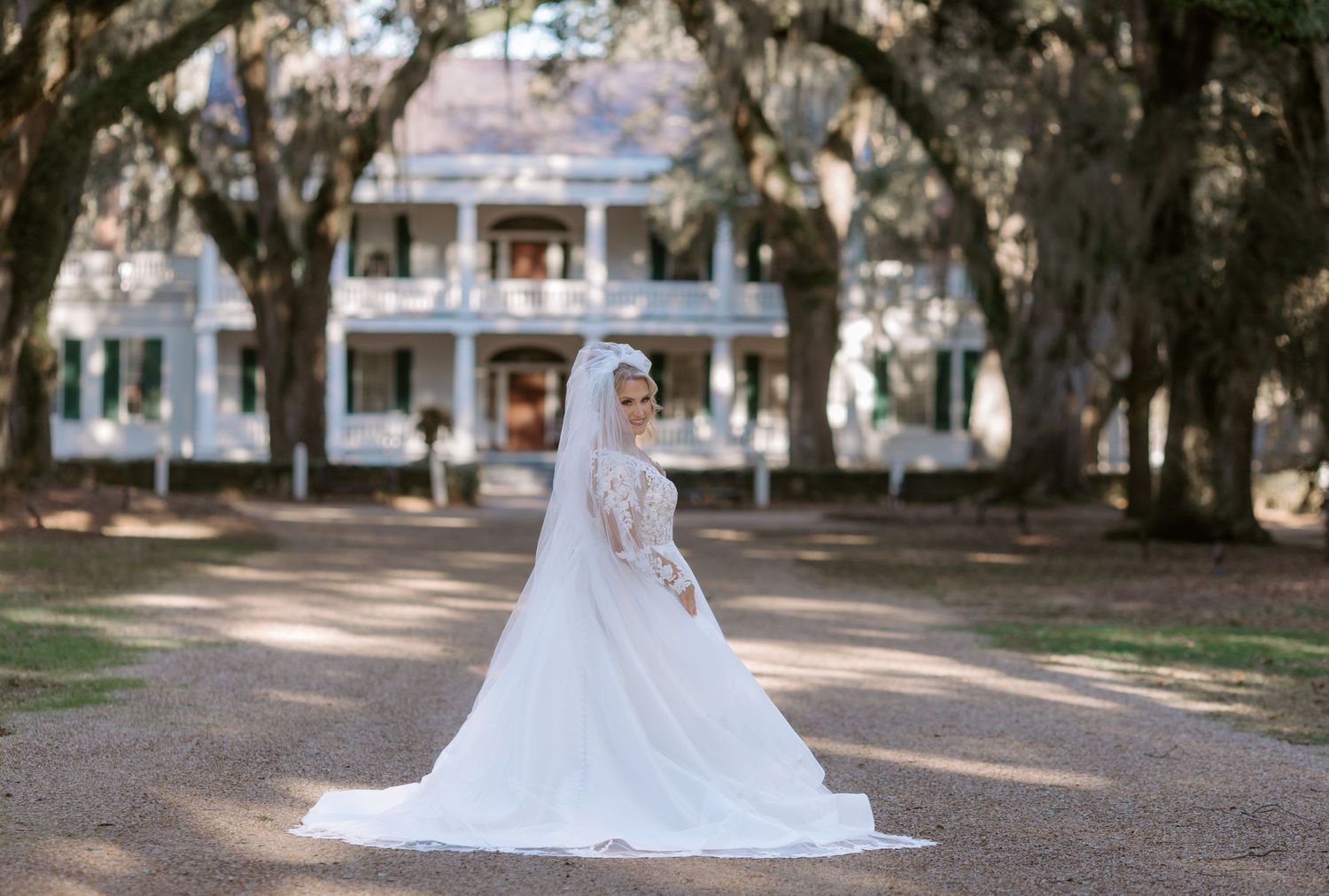 A bride in a flowing white lace wedding gown walks along a tree-lined path during an outdoor photo session.