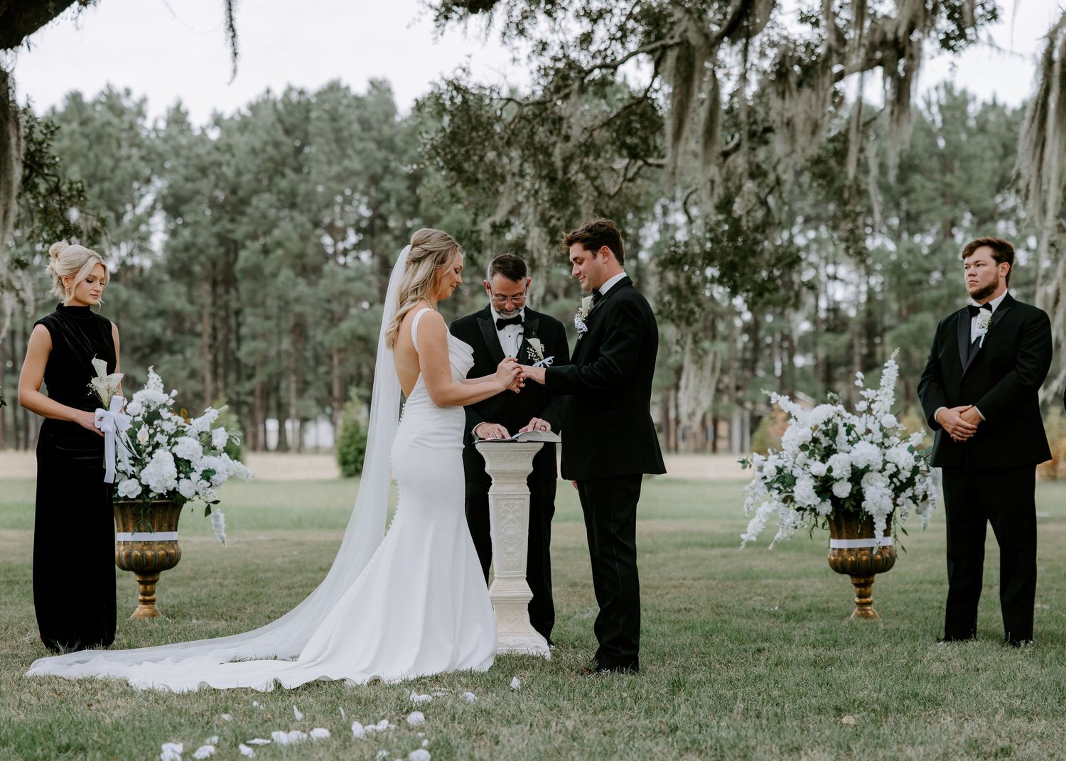Bride and groom exchange vows during an outdoor wedding ceremony beneath Spanish moss-draped oaks, with an officiant and guests looking on.