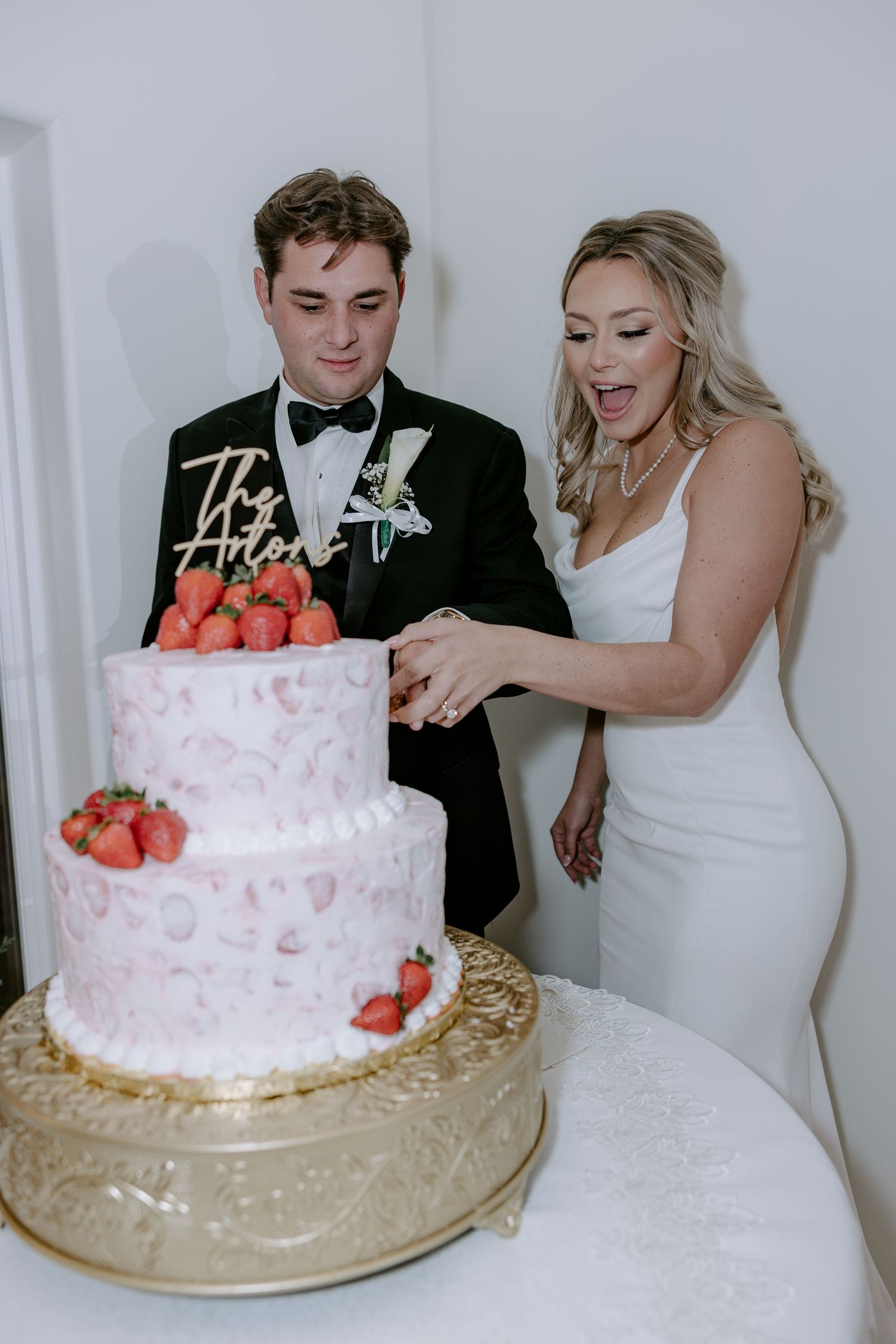Bride in a white gown and groom in a black tuxedo cut a pink wedding cake topped with fresh strawberries on a gold cake stand.