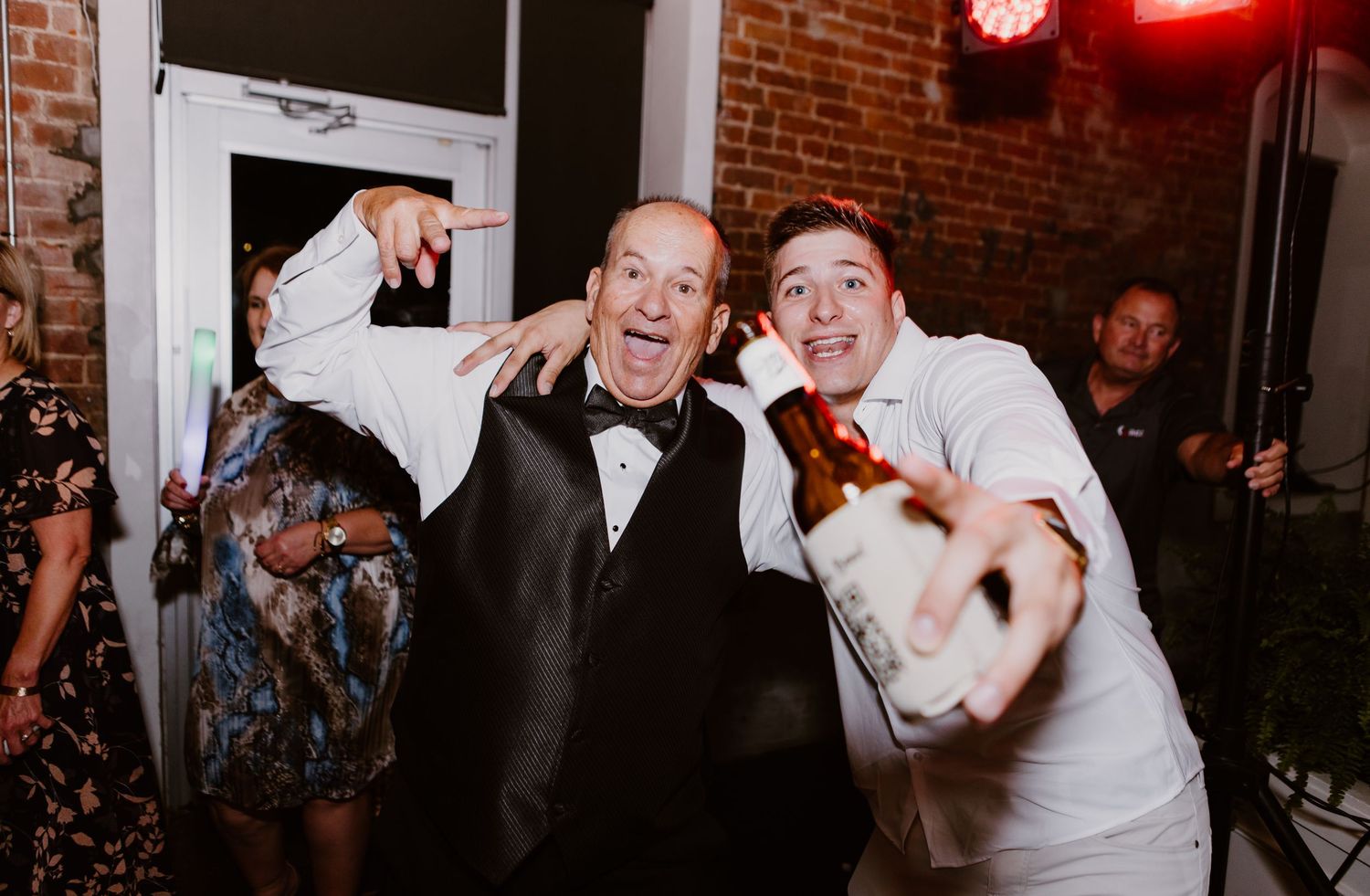 Two men in formal attire pose and celebrate at a lively wedding reception party.