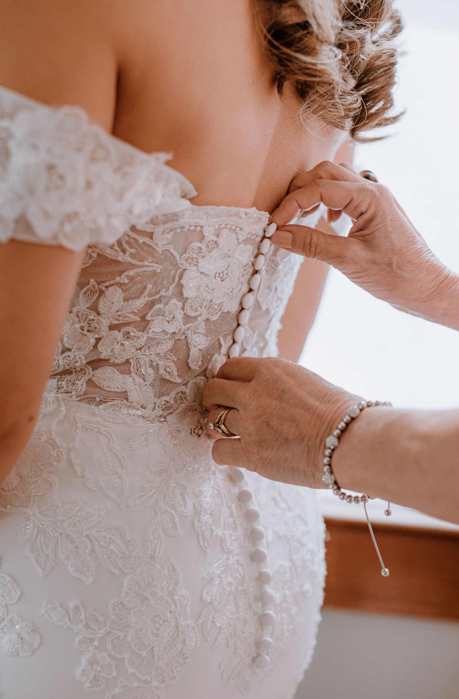 Hands fastening pearl buttons on the back of a white lace off-shoulder wedding dress.