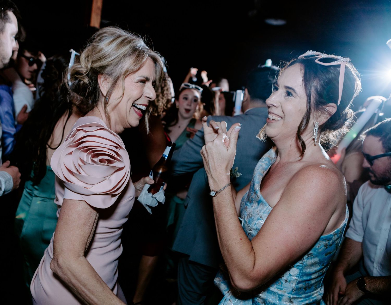 Two women in elegant dresses laughing and dancing together at a festive wedding reception celebration.
