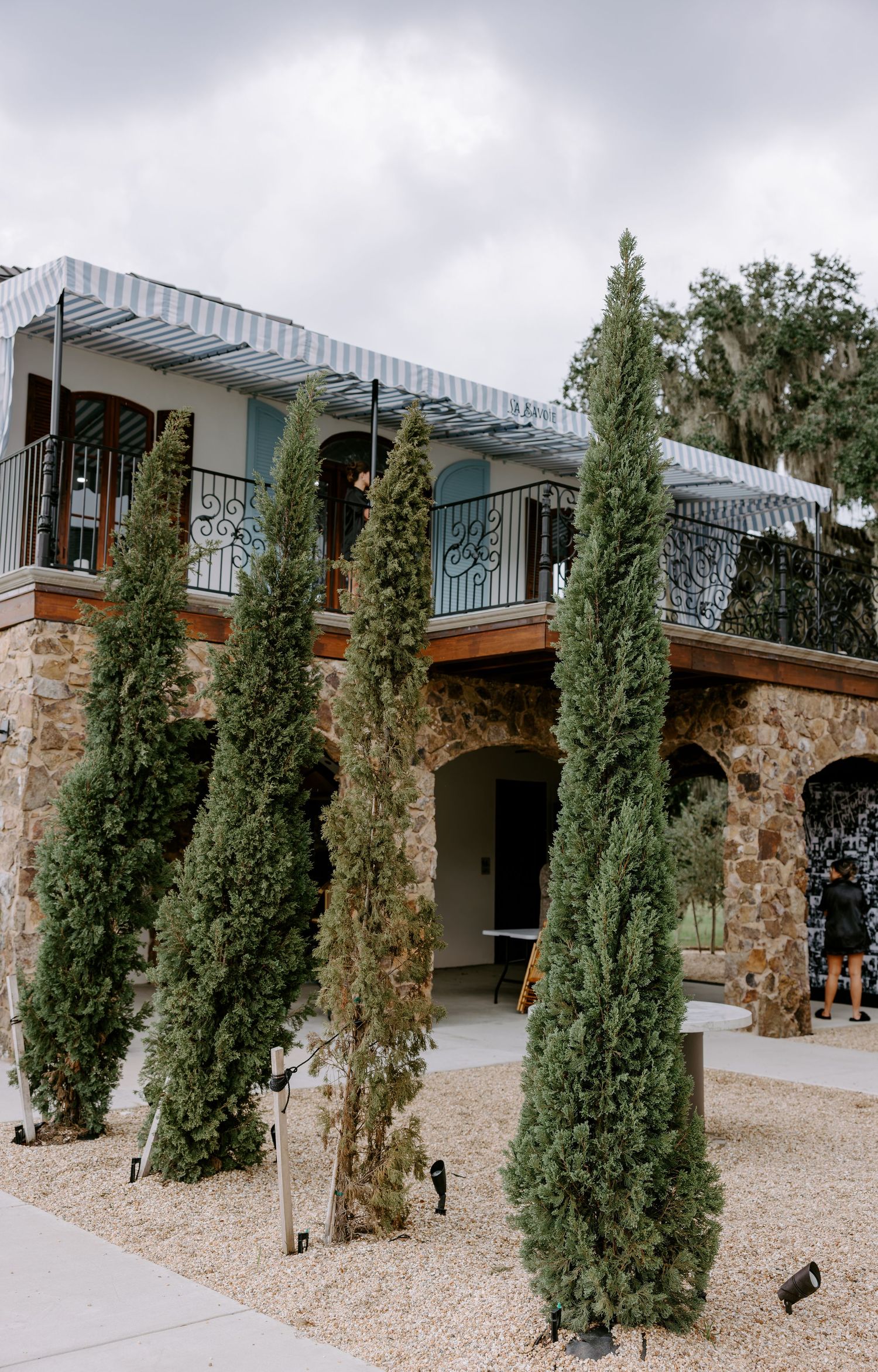 Tall Italian cypress trees line the gravel courtyard of a two-story stone venue with arched doorways and a wrought iron balcony.