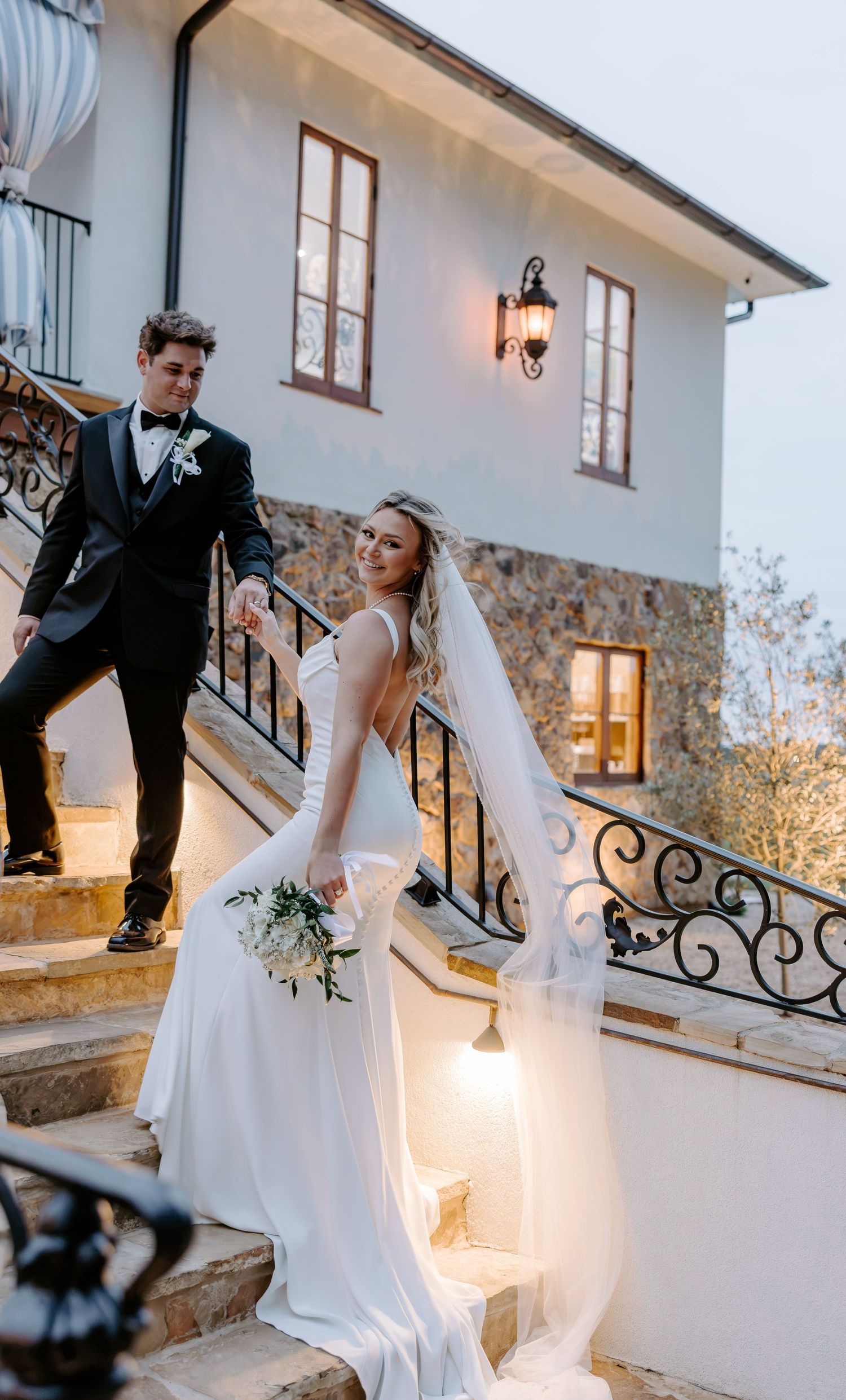 Bride in a white gown with a flowing veil holds a bouquet while walking up stone steps with a groom in a black tuxedo at an elegant venue.