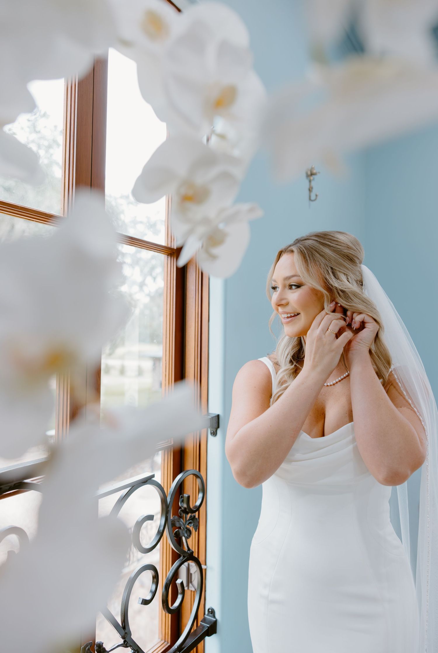 Smiling bride in an elegant white gown and veil adjusts her earring by a sunlit window, framed by white orchids in a soft blue room.