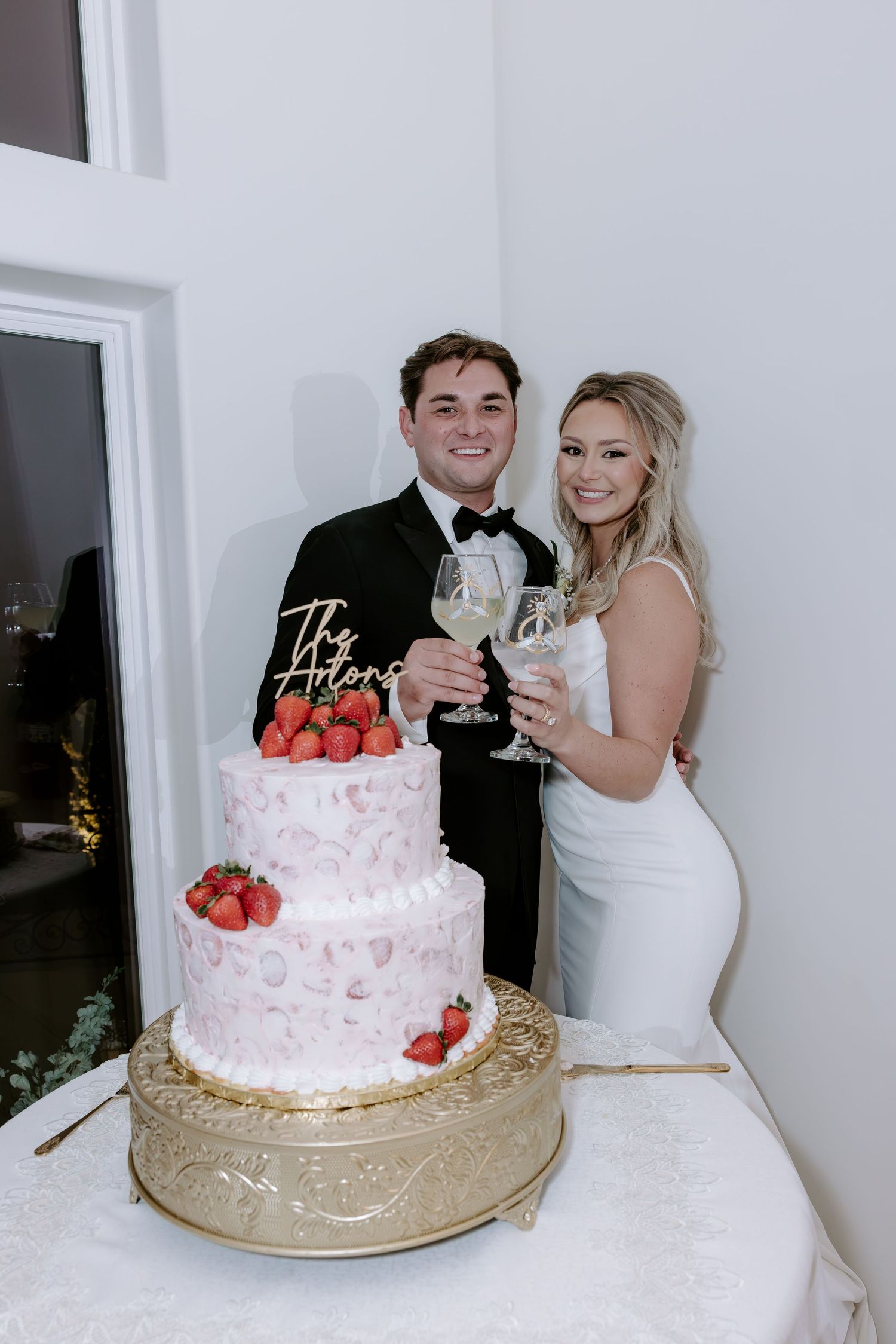 Smiling newlywed couple in a black tuxedo and white gown holding champagne glasses beside a pink strawberry wedding cake on a gold stand.