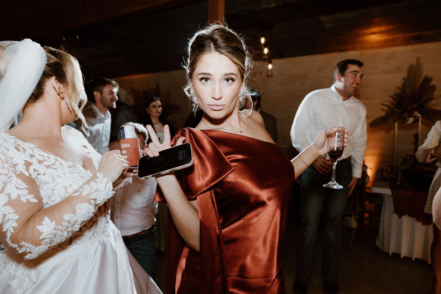 Bridesmaid in rust satin off-shoulder gown holds drink at elegant fall wedding reception.