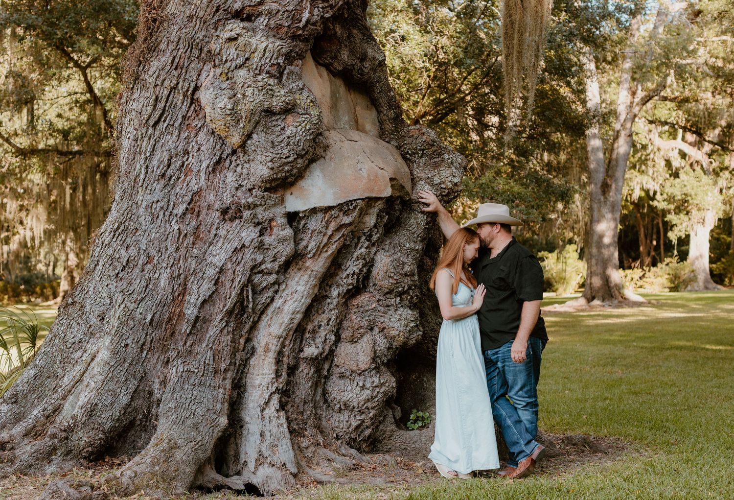 A couple in blue and denim poses for romantic photos among large oak trees in a sunlit park setting.