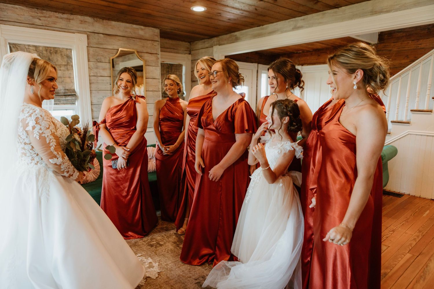 Bridesmaids in rust red gowns admire the bride in a white lace wedding dress with a young flower girl.