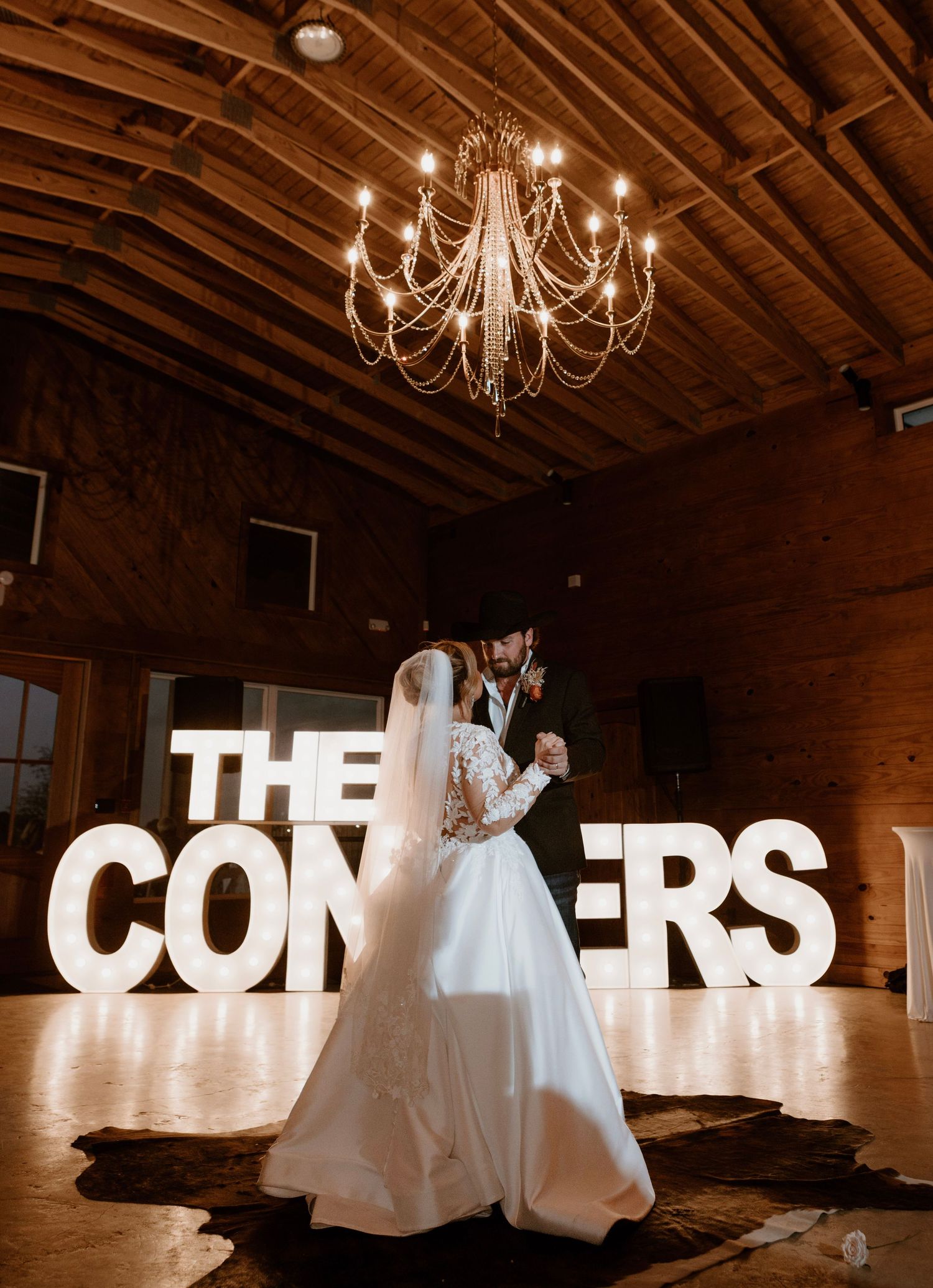 Couple dancing beneath elegant chandelier in front of glowing THE CONNERS marquee letters in a rustic barn wedding venue.