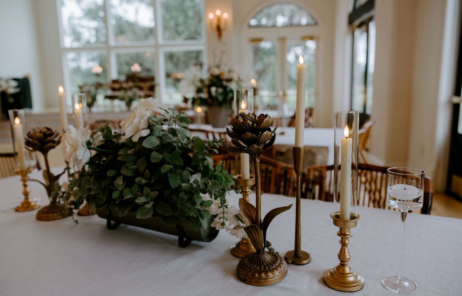 Elegant wedding reception table with greenery centerpiece, gold candlesticks, and wine glasses in a bright room with tall arched windows.