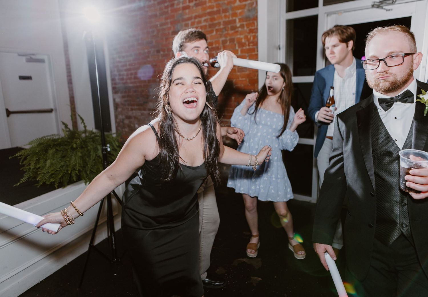 Guests laughing and dancing at a wedding reception, woman in black dress smiling, man in tuxedo holding drink.