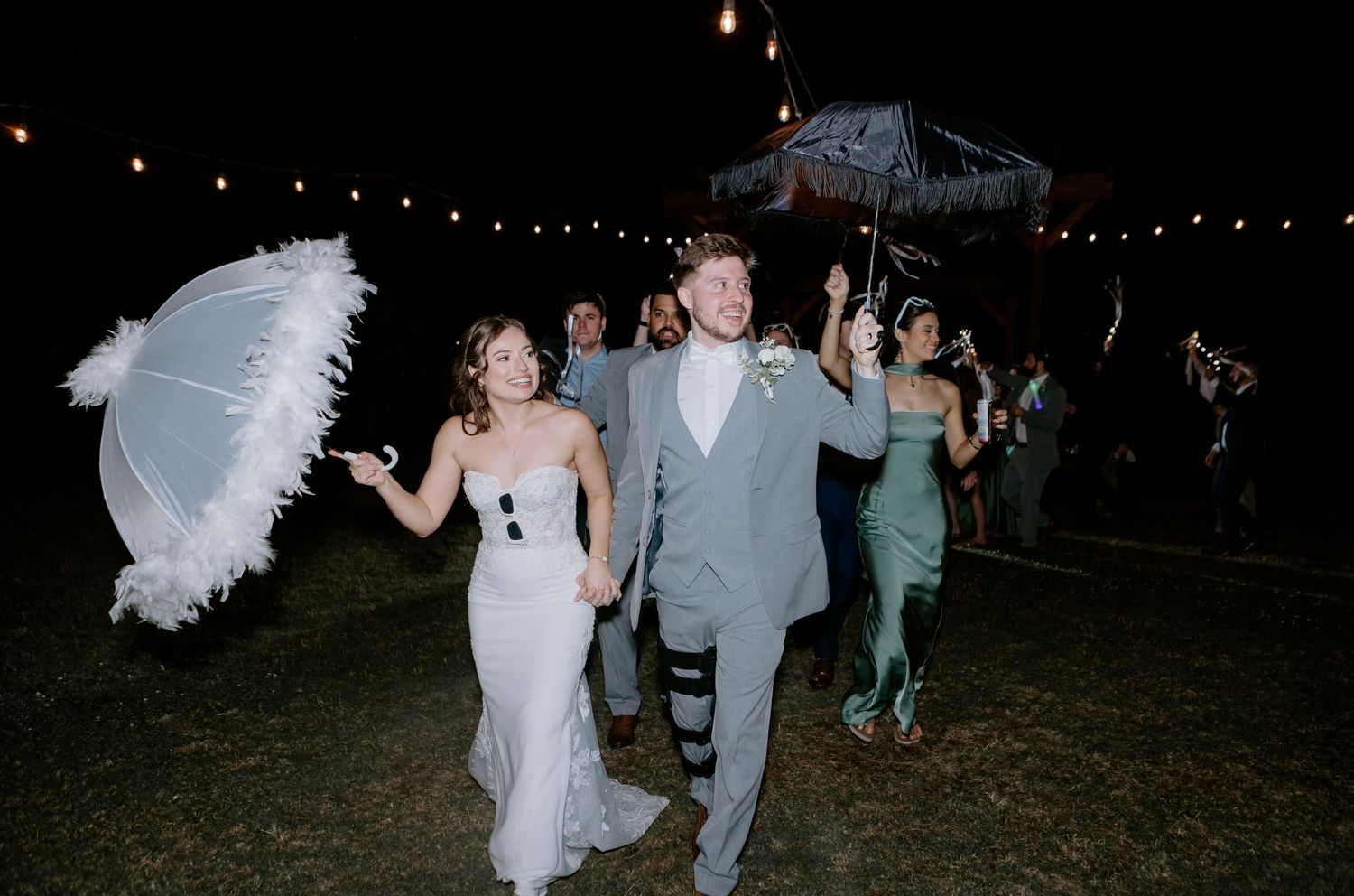 Bride and groom exiting wedding reception at night carrying feathered umbrellas under string lights.