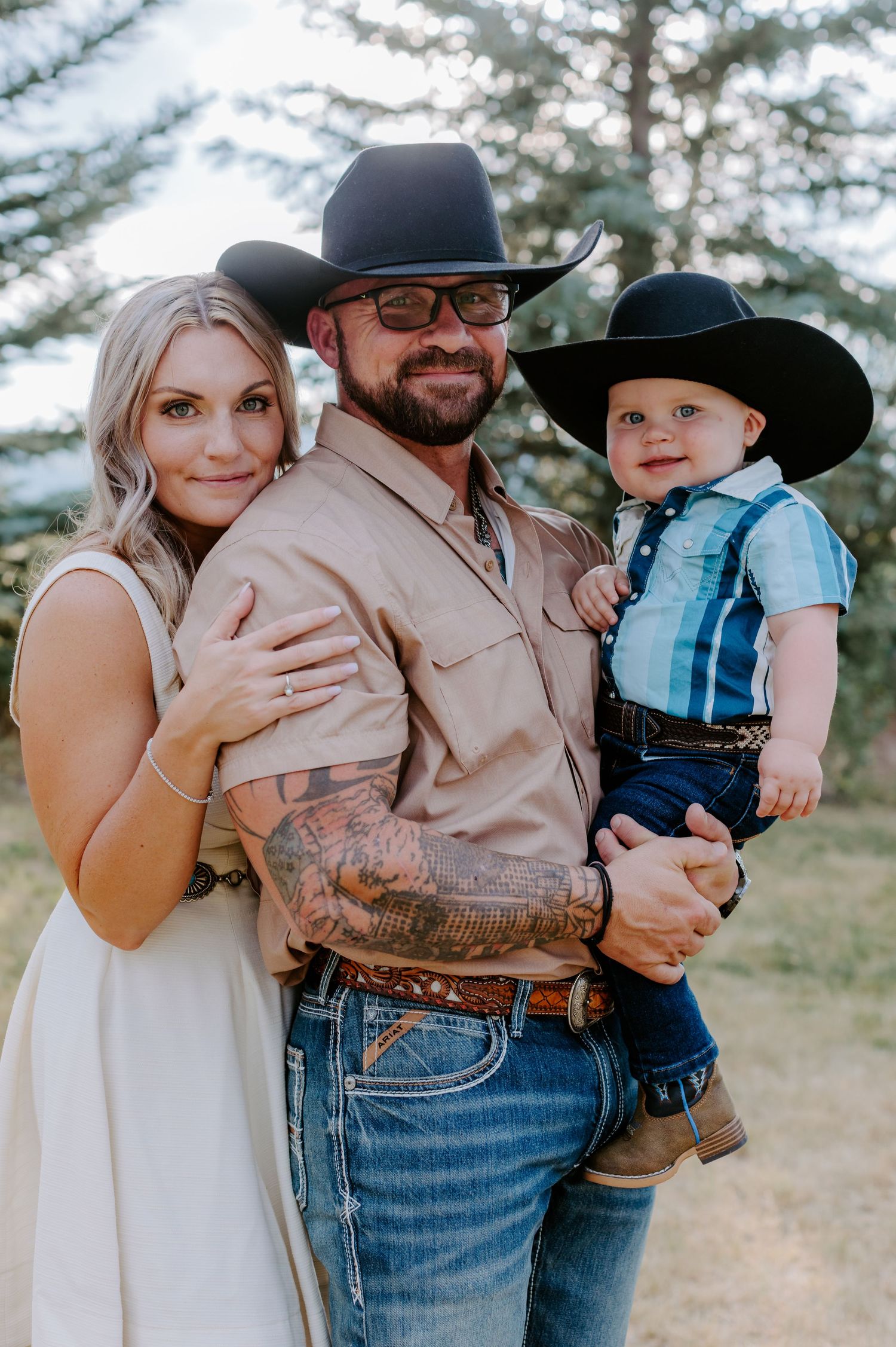 A family of three in Western attire, with cowboy hats, posing outdoors near pine trees.