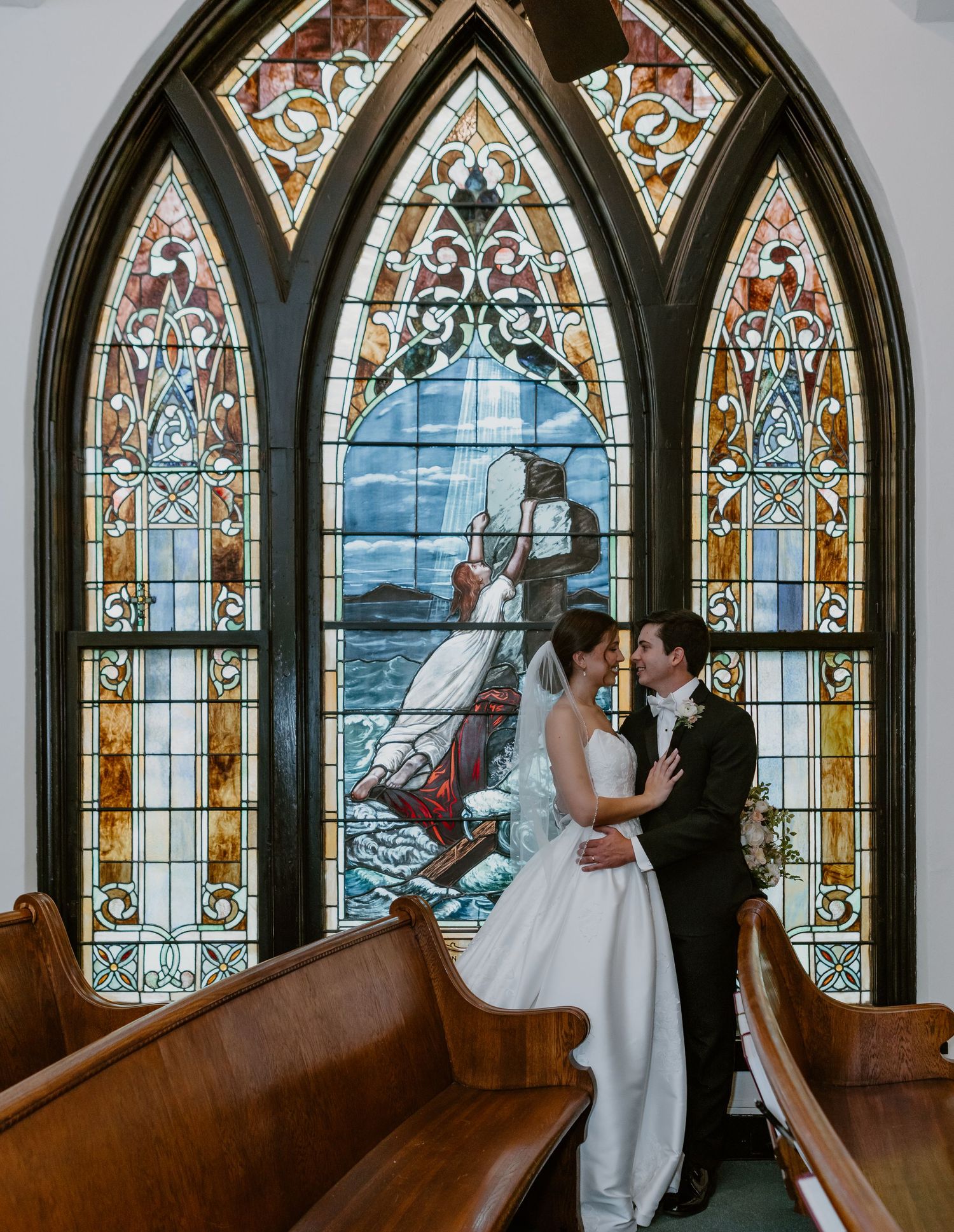Bride and groom share a kiss in front of a stunning stained glass window inside a historic church.