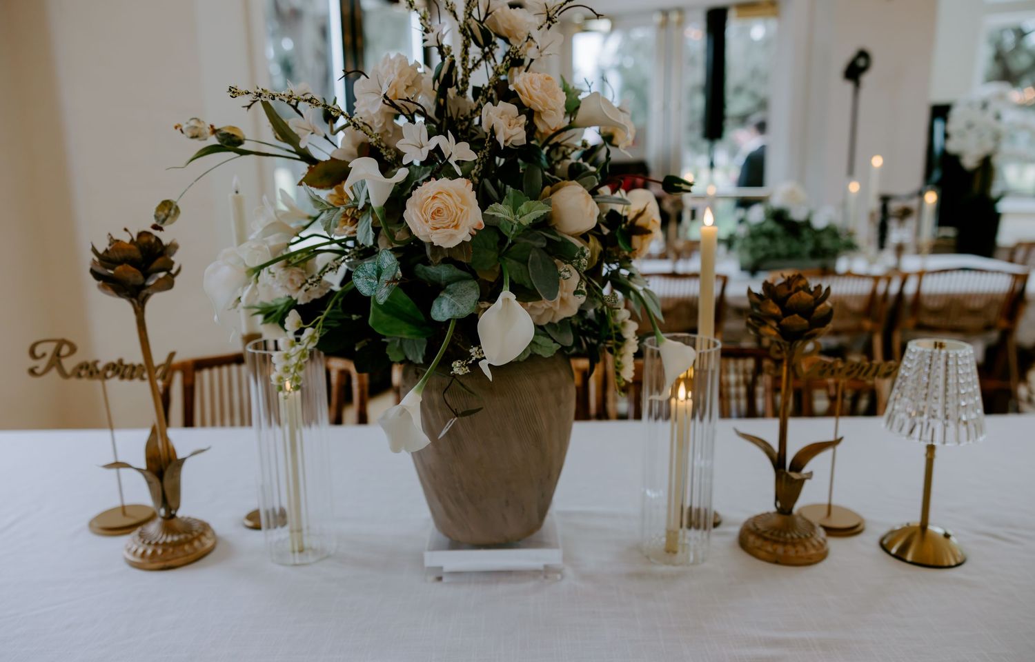 Elegant wedding table centerpiece with cream roses and greenery in a stone vase, surrounded by gold candle holders and lit taper candles.