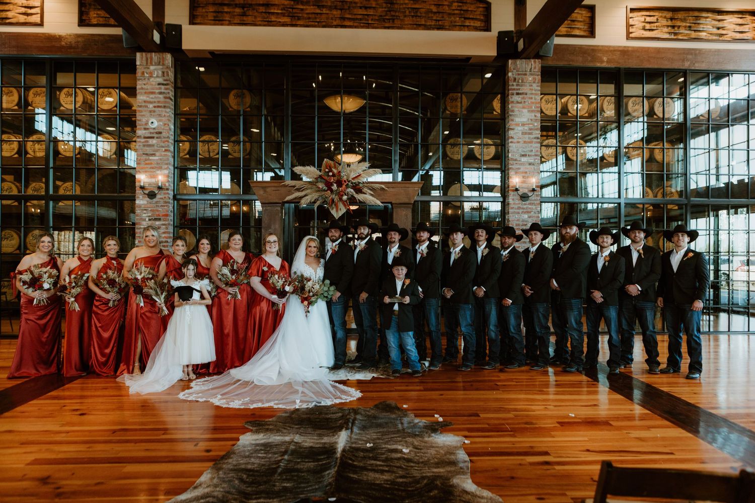 Wedding party poses in rustic lodge venue, bridesmaids in red gowns, groomsmen in dark suits with cowboy hats.