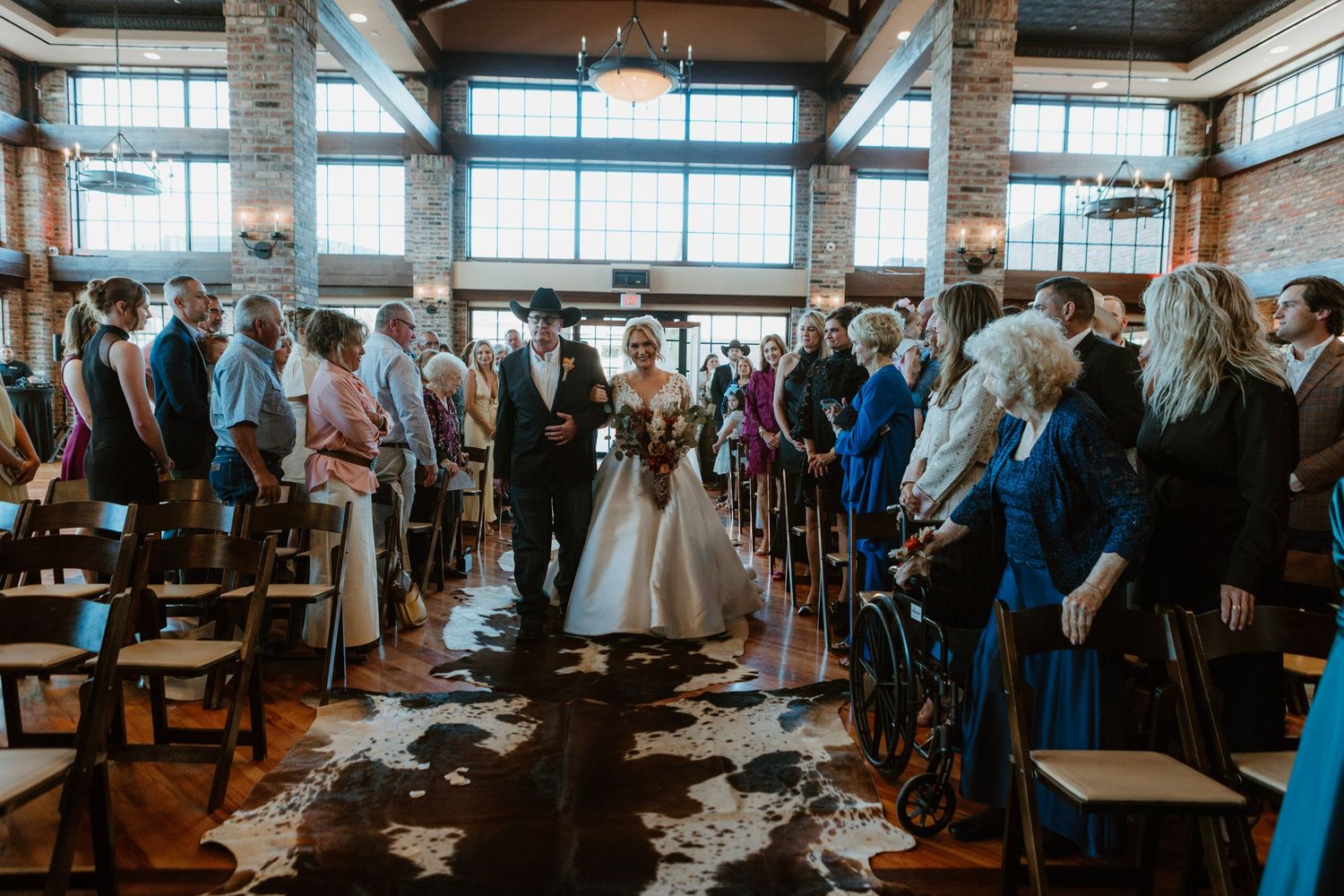 Bride and groom walk down the aisle at an elegant indoor wedding ceremony with guests standing in a rustic venue.