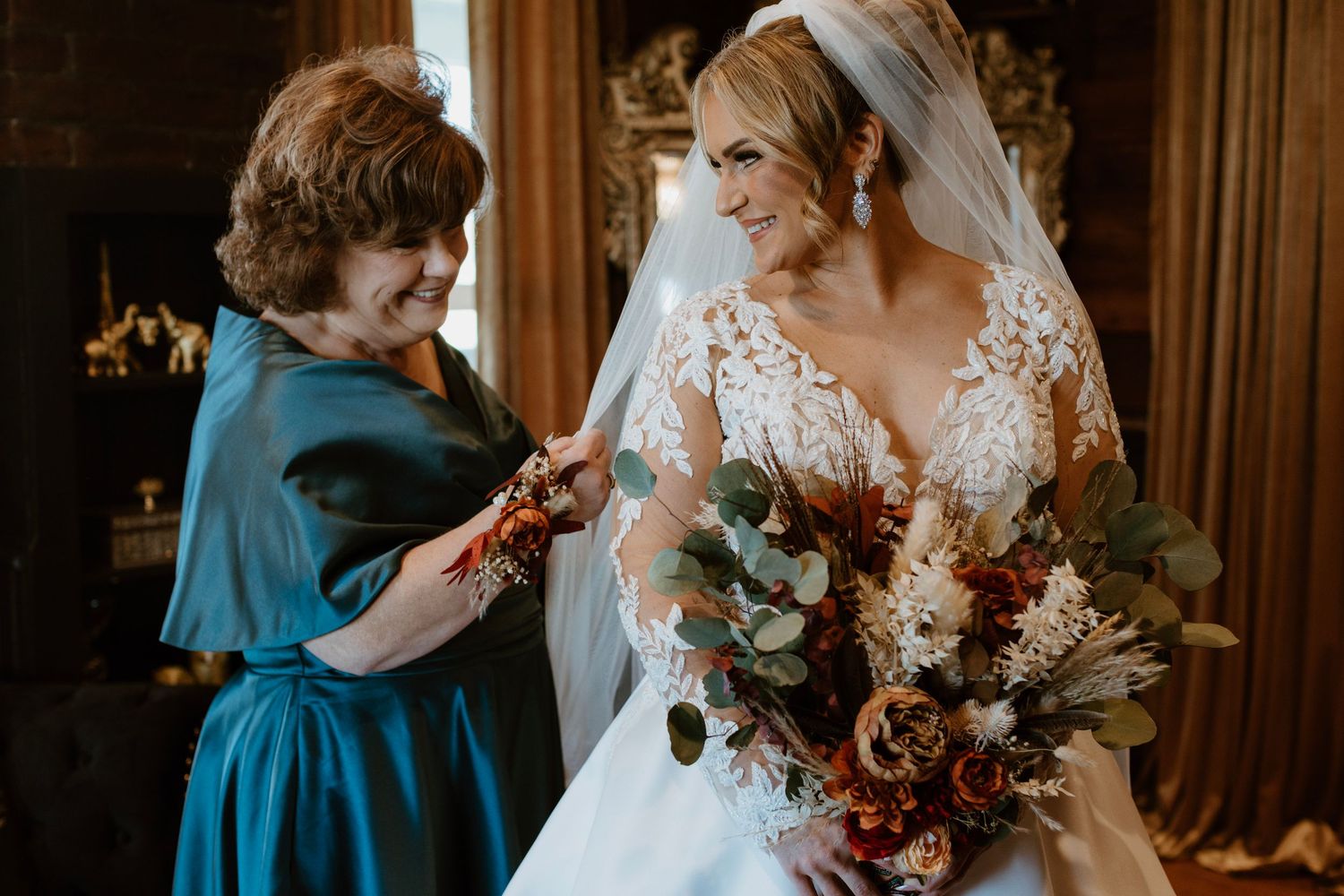 Bride in lace wedding gown holding autumn bouquet with older woman in teal dress assisting during wedding preparations.