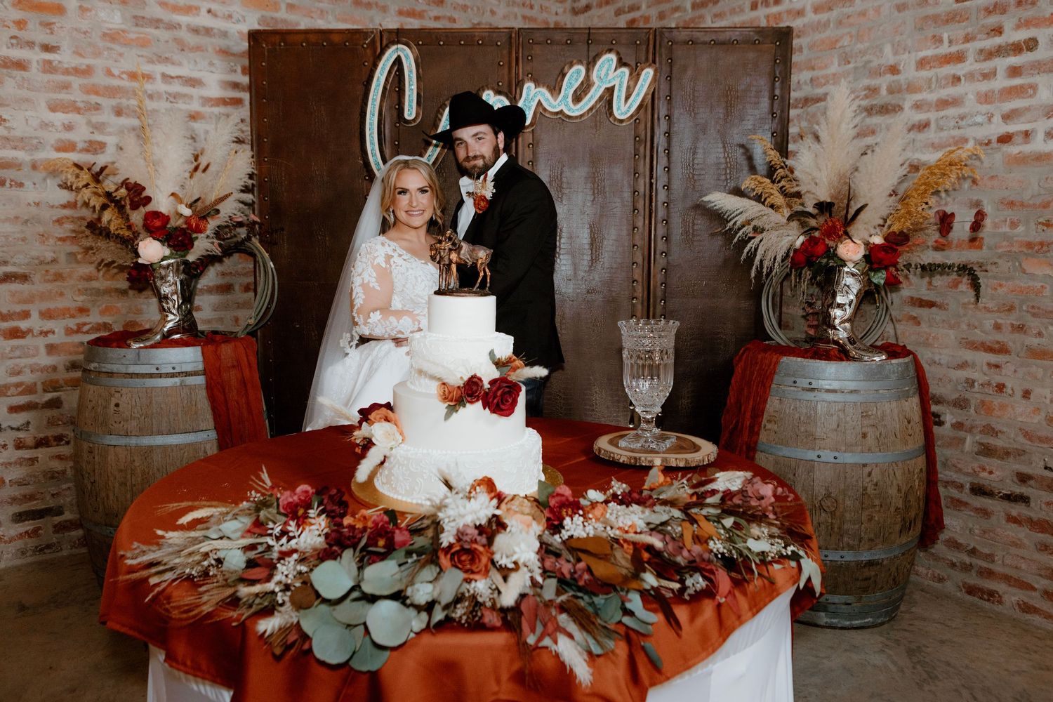 Newlyweds cut white wedding cake at decorated table with fall florals and wine barrels at rustic venue.