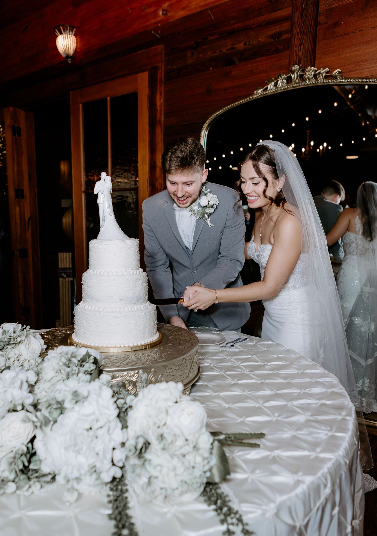Bride and groom smiling while cutting their white tiered wedding cake decorated with floral topper.