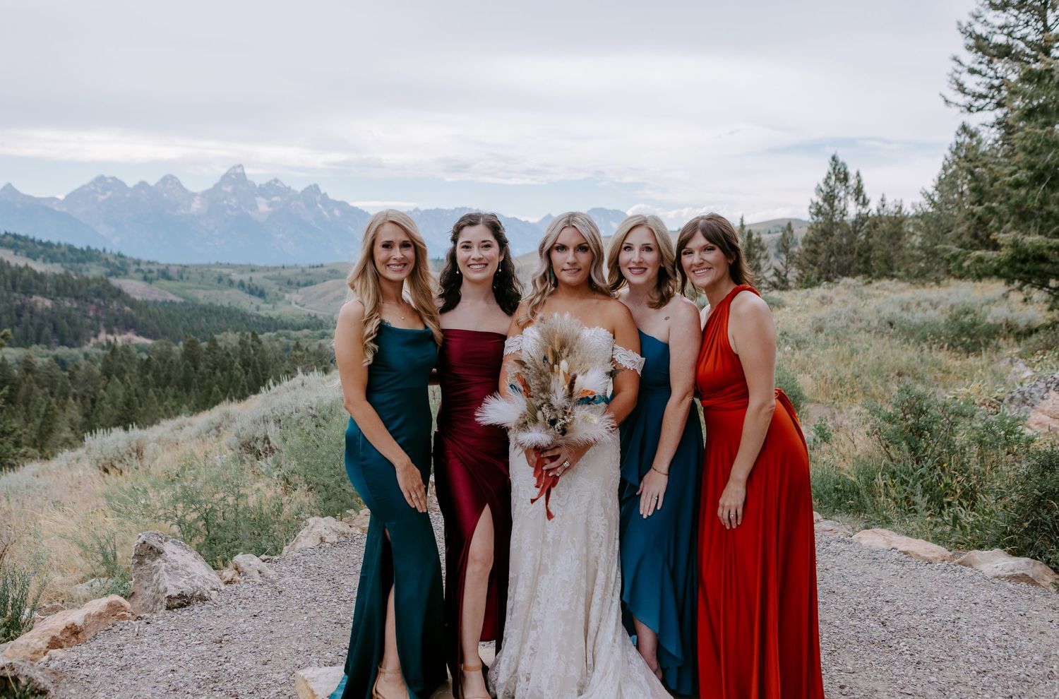 Bride poses with four bridesmaids in red and teal gowns with stunning Grand Teton mountain views behind them.