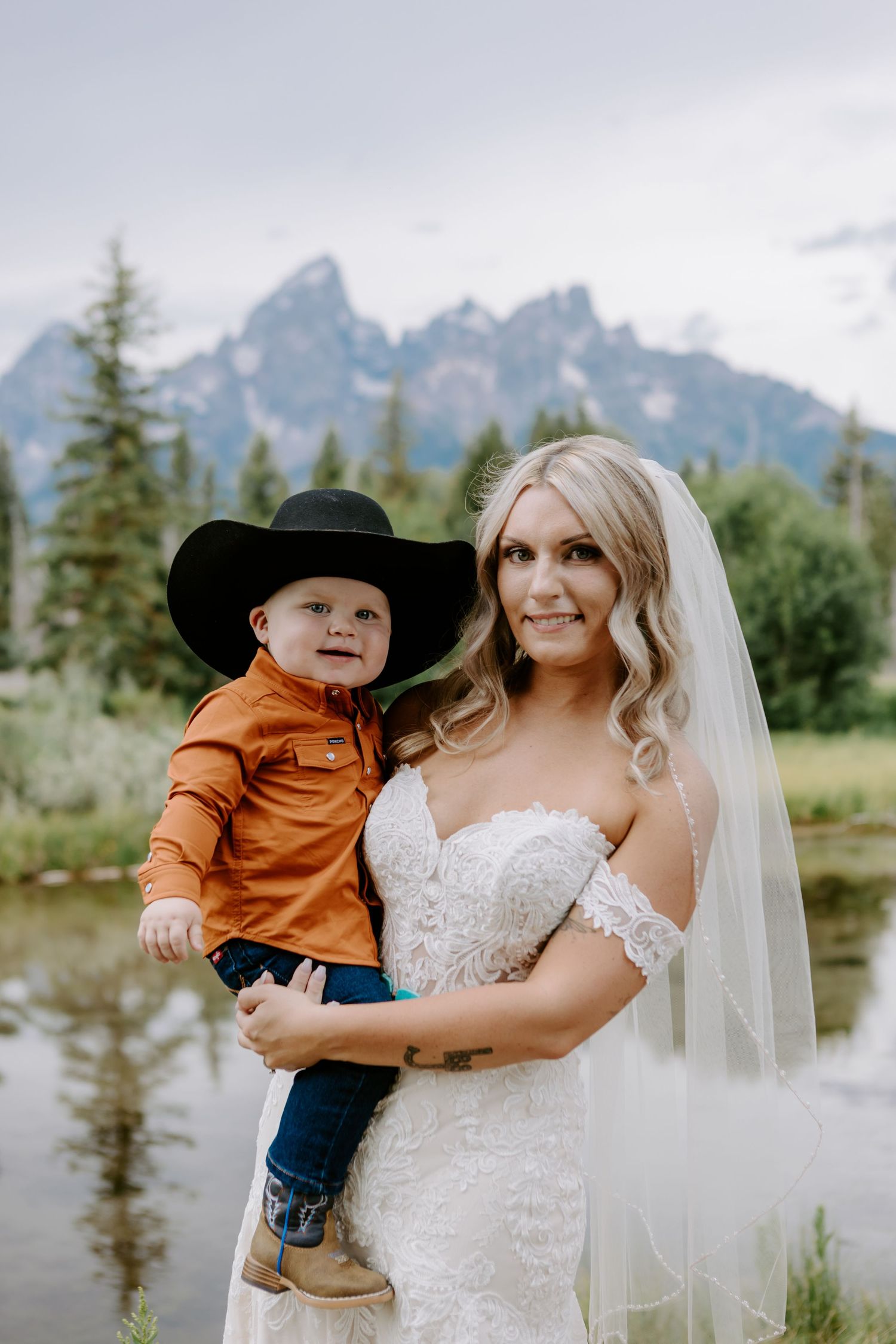 Bride in lace off-shoulder gown holding young boy in cowboy hat with Grand Teton mountains backdrop.