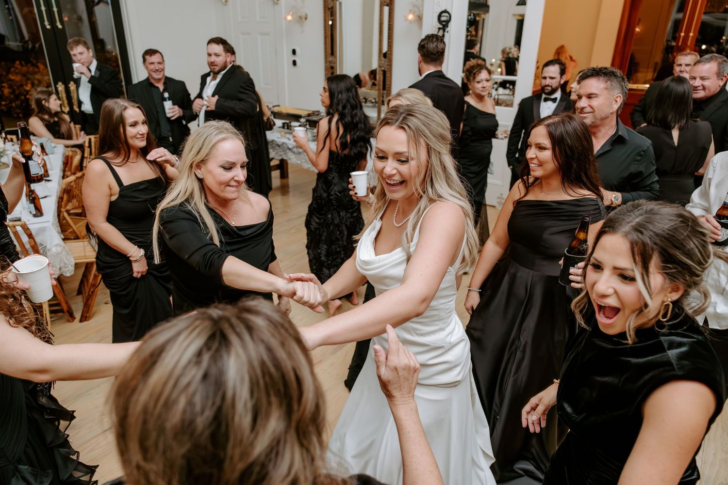 Bride in a white dress dances joyfully with smiling guests in black dresses during a lively wedding reception.