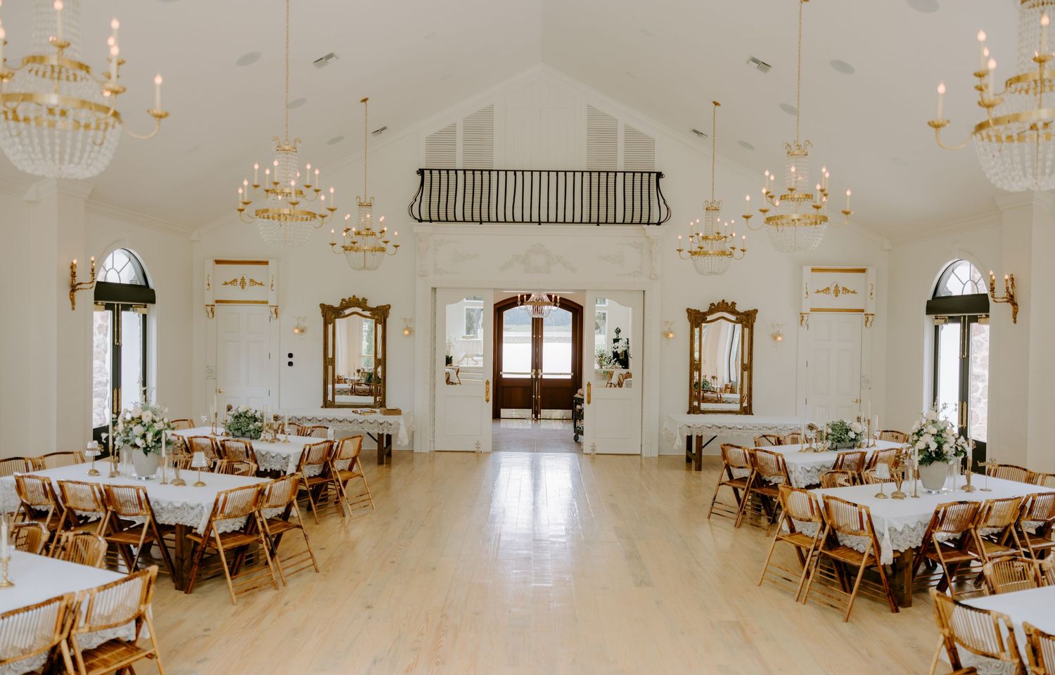 Elegant wedding venue ballroom with crystal chandeliers, wooden floors, arched windows, and rows of banquet tables with wooden chairs.