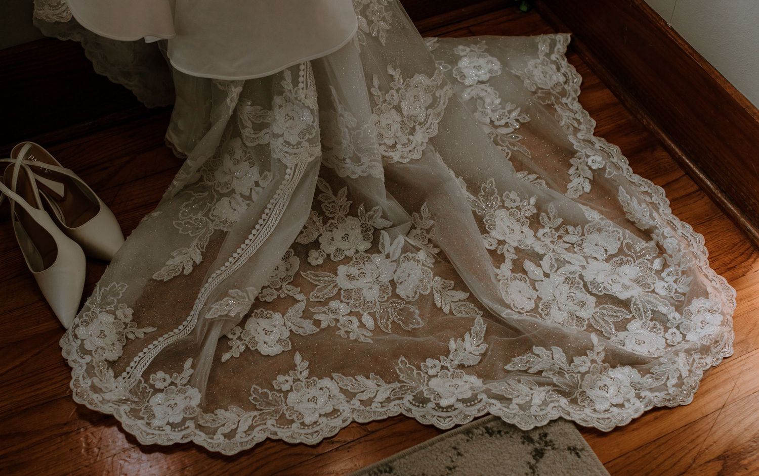Elegant lace wedding dress train with floral embroidery spread on hardwood floor beside white bridal heels.
