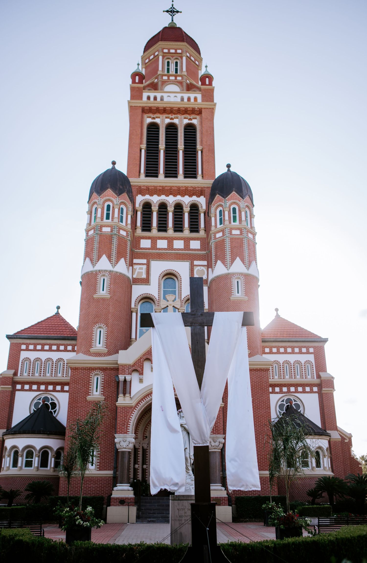 Red brick cathedral with bell tower and large decorative M cross display in foreground under clear sky.