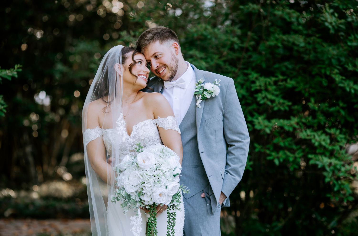 Bride and groom smiling together outdoors, bride holding white floral bouquet wearing veil and off-shoulder gown.