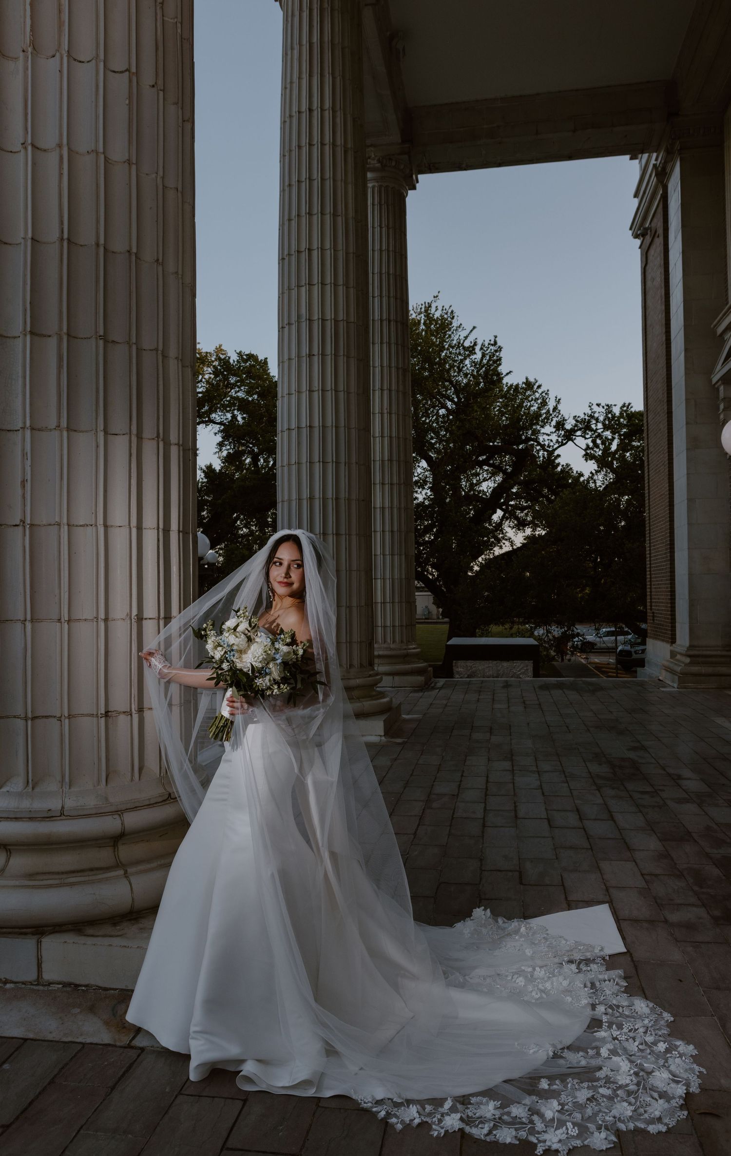 A bride in a flowing white wedding gown and veil stands elegantly between classical stone columns at dusk.