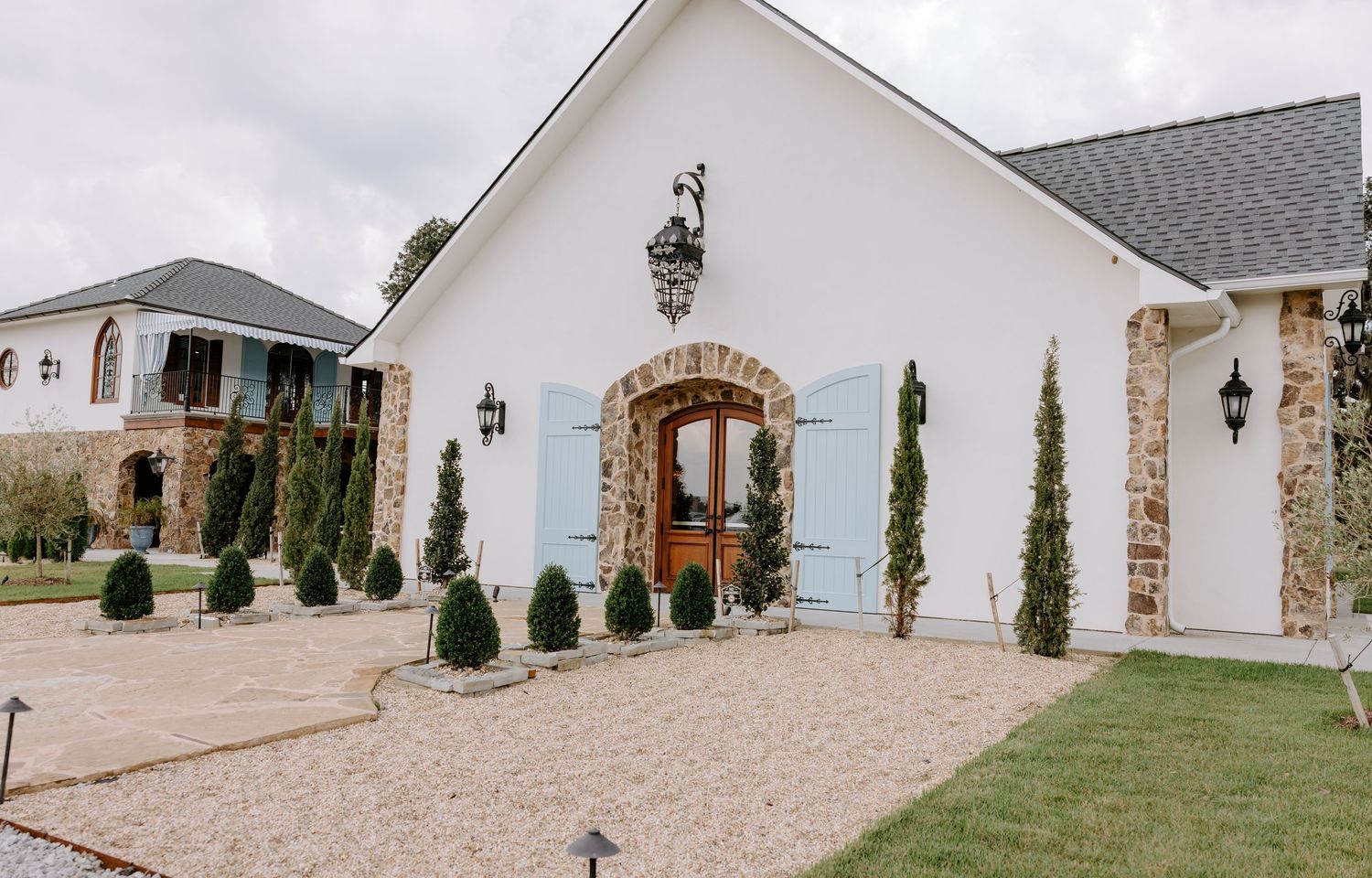 White building with stone arched entry, light blue shutters, wall lanterns, and manicured shrubs along a gravel walkway.