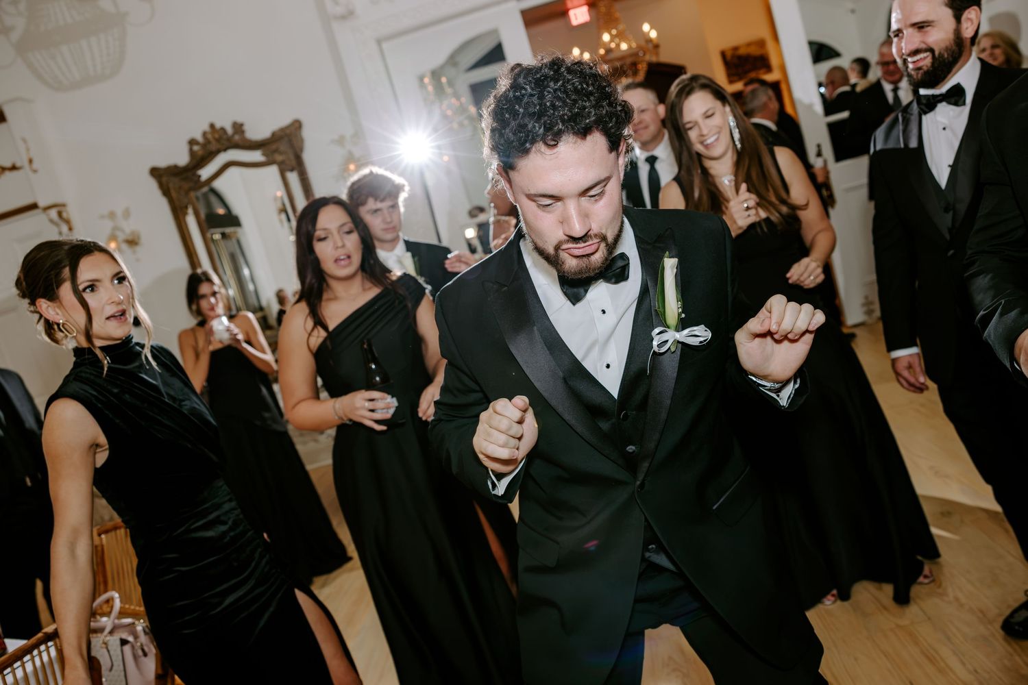 Bearded groom in a black tuxedo dances joyfully on the dance floor while elegantly dressed wedding guests look on..