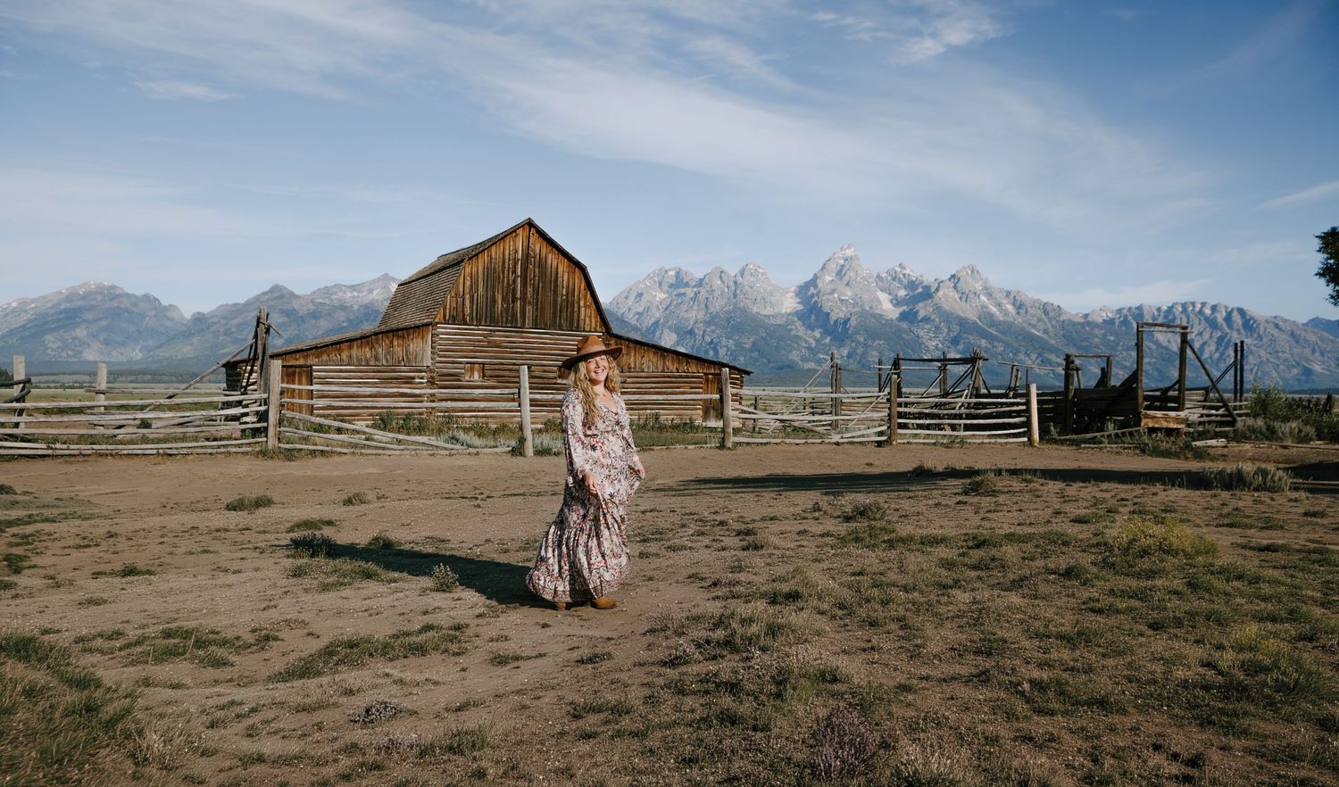Woman in floral maxi dress walks near historic Mormon Row barn with Grand Teton mountains backdrop.
