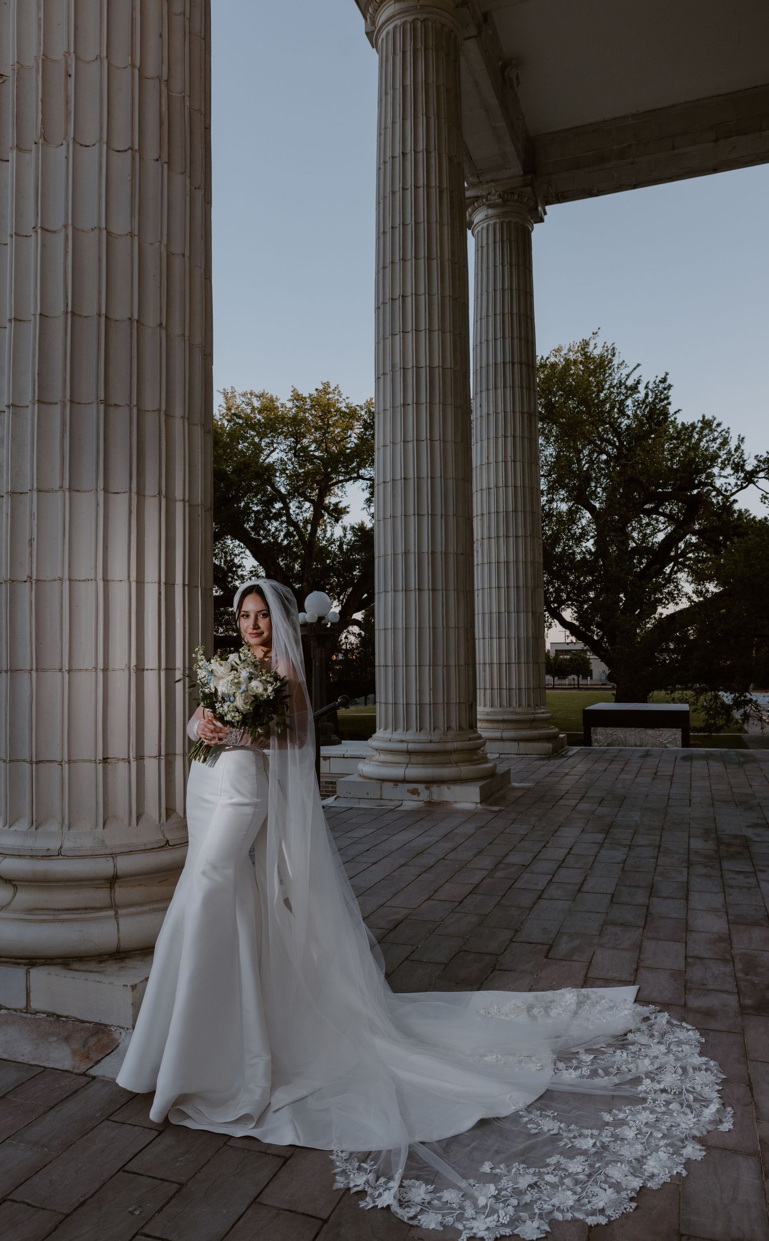 An elegant white bridal gown with a flowing lace veil stands between classic stone columns at a grand venue.