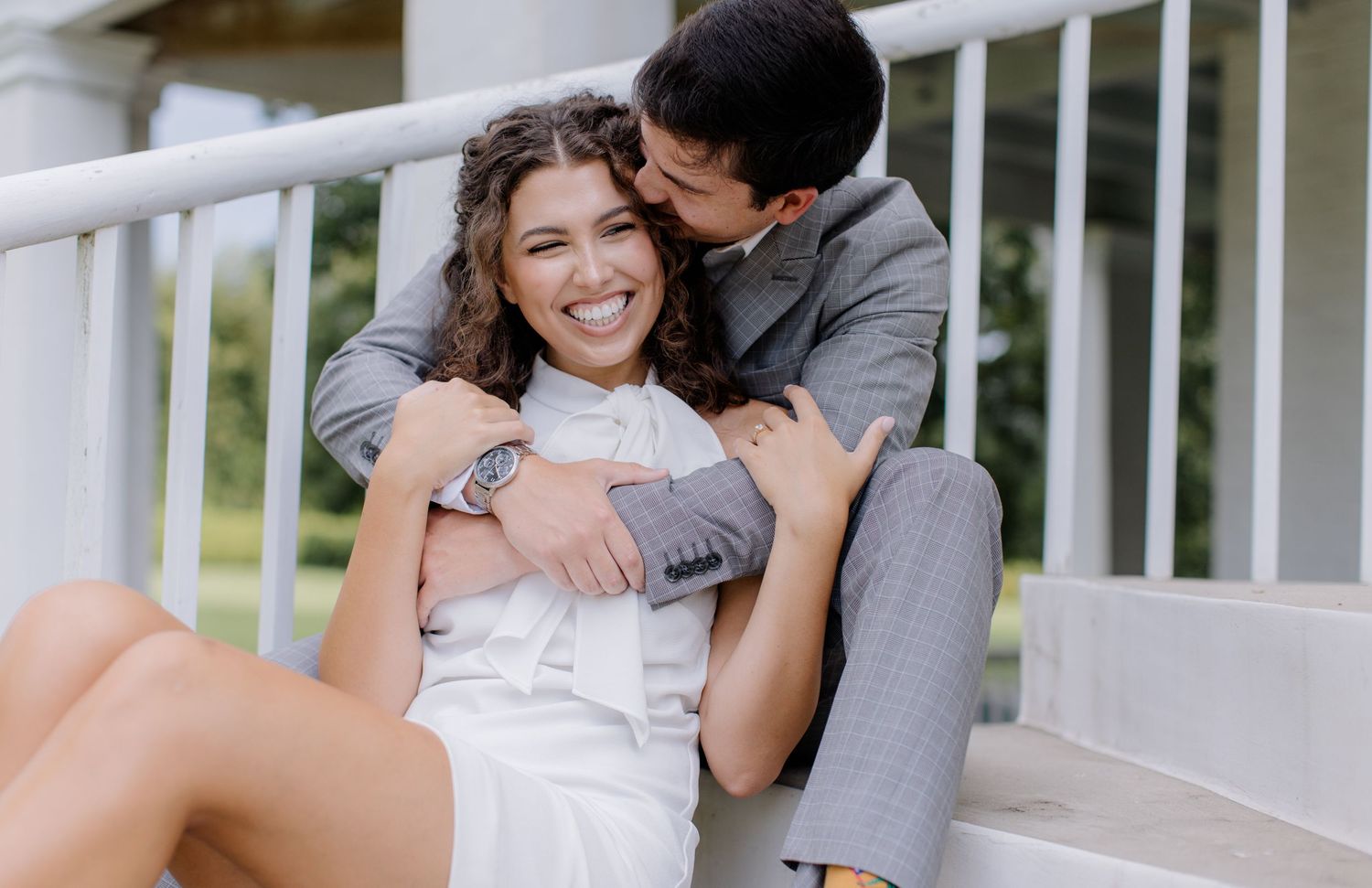 A couple shares a tender moment while sitting on white porch stairs, wearing gray and white outfits.