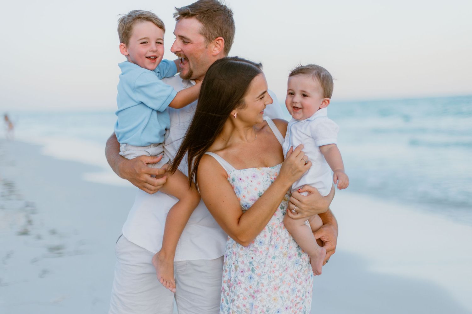 A family shares joyful moments together during a photoshoot on a sunny beach with ocean waves in the background.