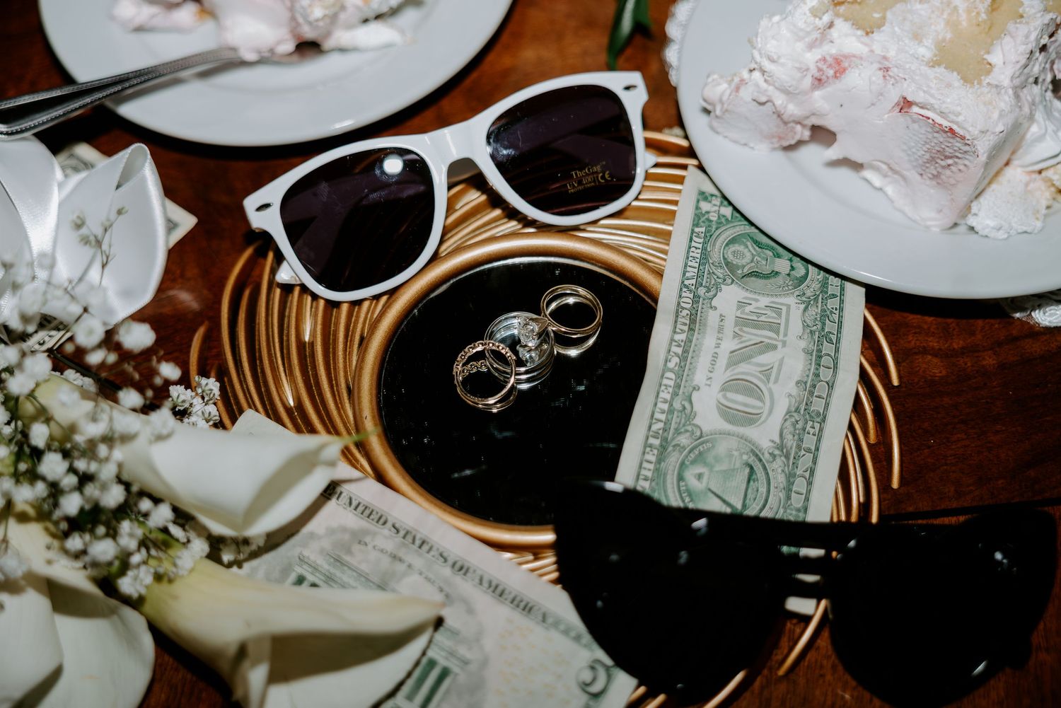 Wedding rings rest on a round gold tray beside white sunglasses, a dollar bill, baby's breath flowers, and napkins on a wooden table.