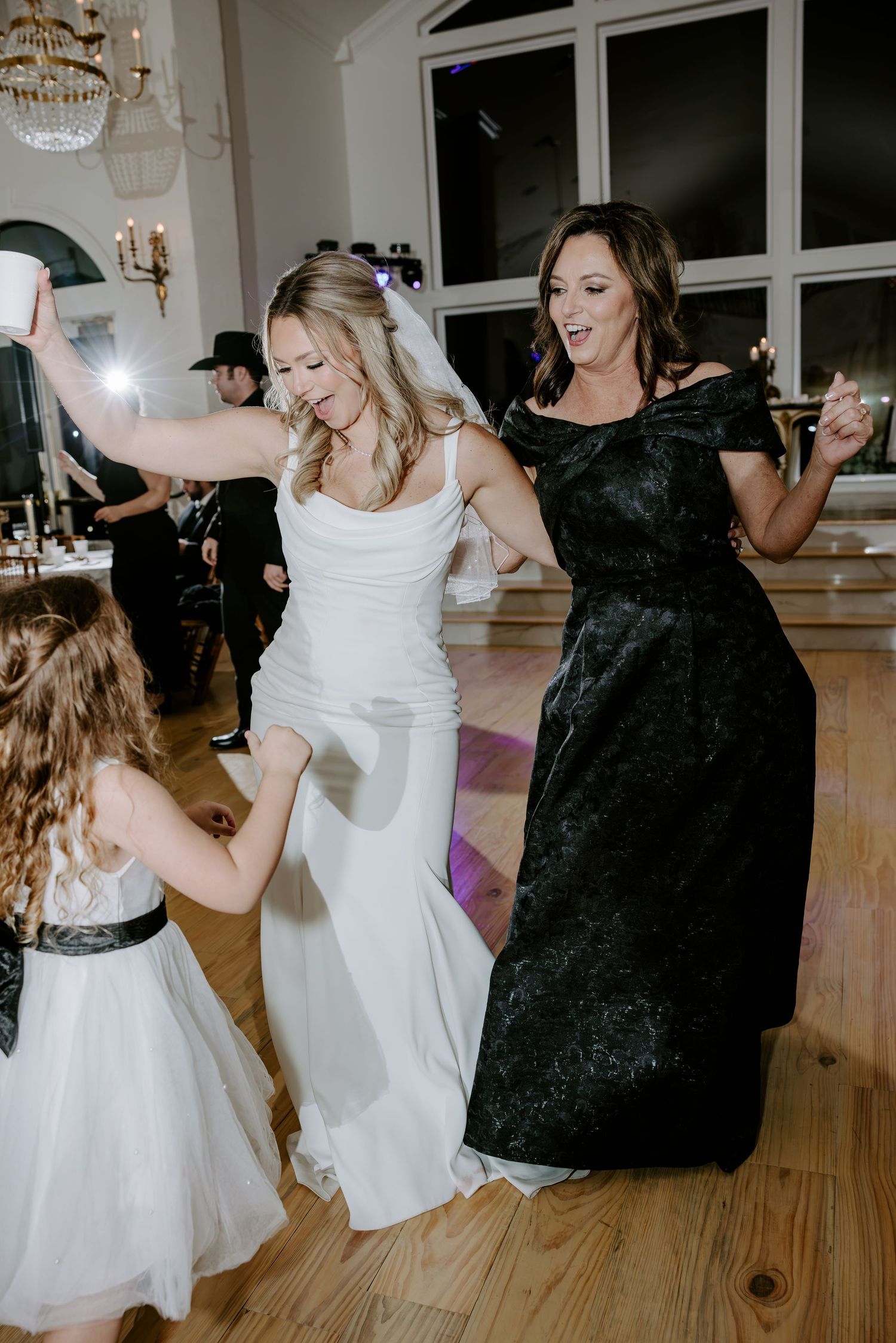 Bride in a white gown and veil dances joyfully with a woman in a black dress and a young flower girl on a wedding reception dance floor.