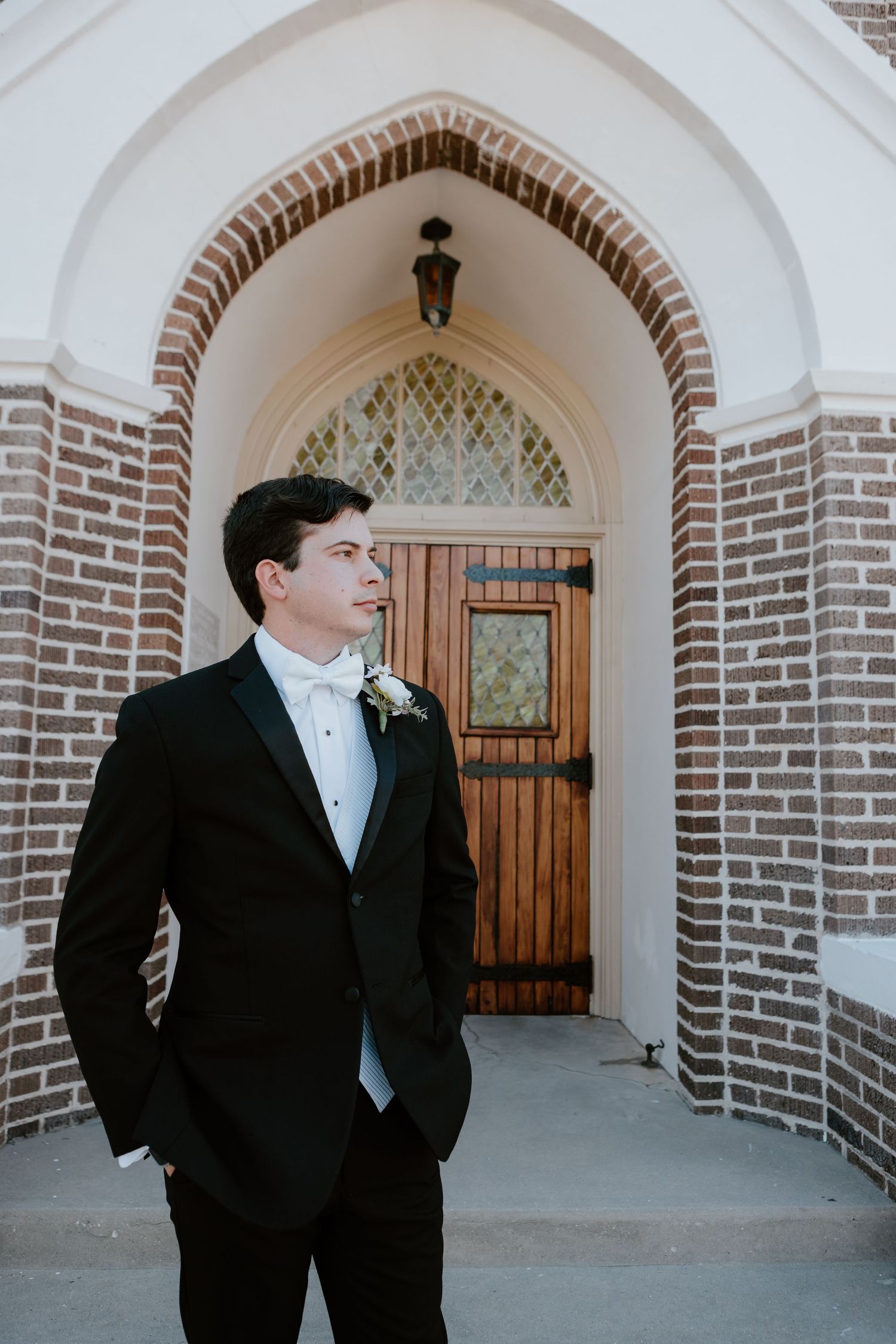 A groom in a black tuxedo with boutonniere stands before a brick church archway, looking thoughtfully to the side.