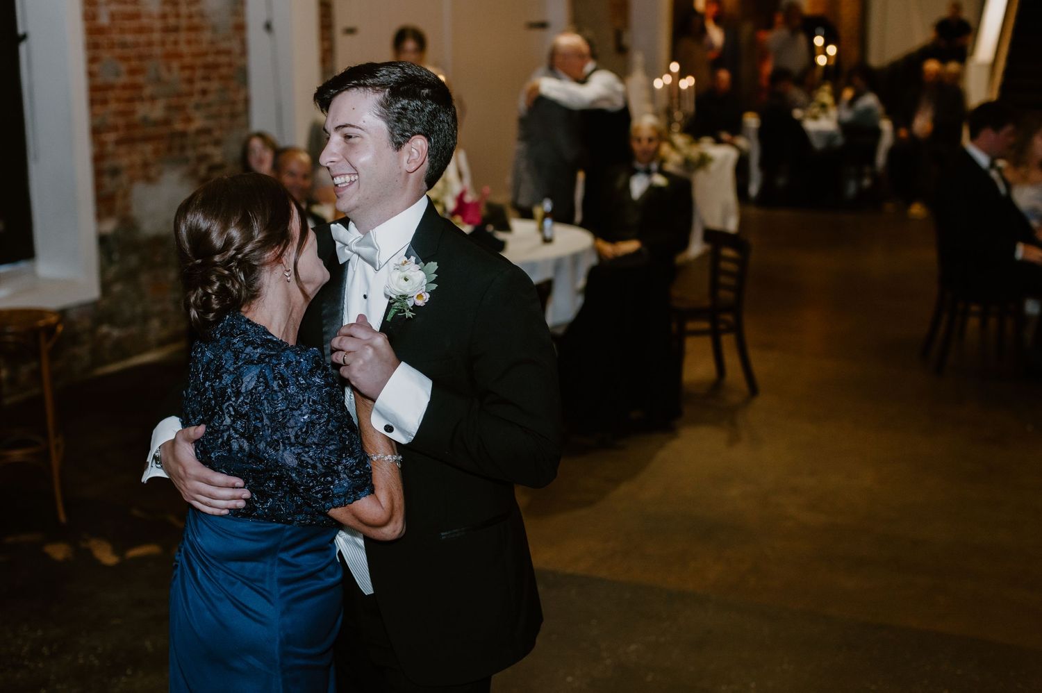 Groom dances with mother in navy dress during mother-son dance at wedding reception.