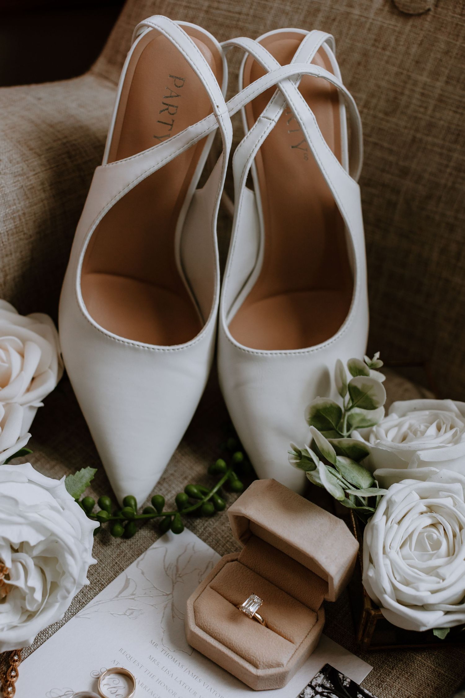 White bridal heels with cross straps beside roses, greenery, and a gold engagement ring in a velvet box.